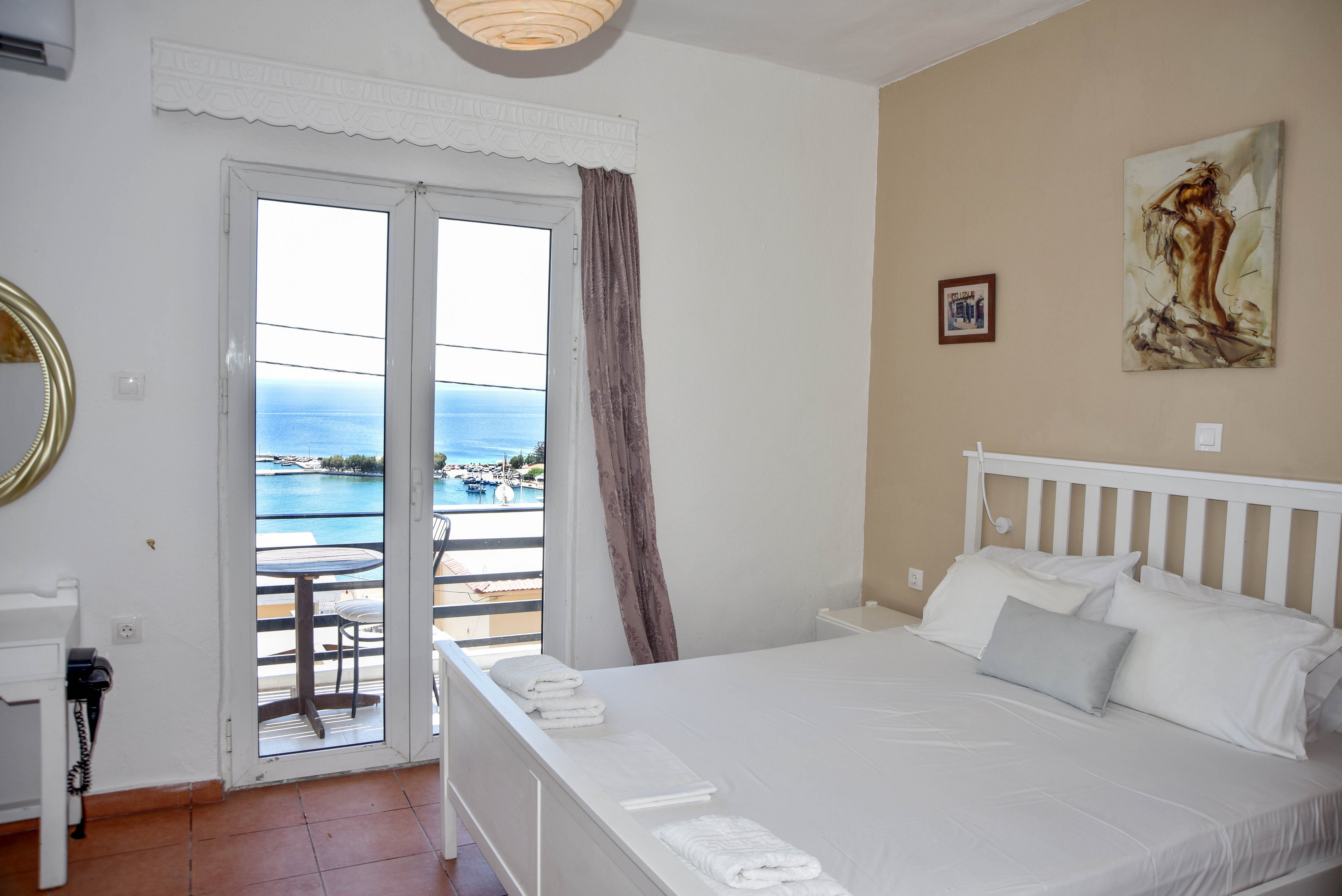 A bright, neatly made double bed dominates a small hotel bedroom with white furniture. French doors open onto a balcony overlooking a calm blue sea and distant coastline. Soft neutral tones, wall art, and folded towels on the bed create a clean and inviting atmosphere. Natural daylight fills the room, emphasizing the relaxed coastal vacation setting.