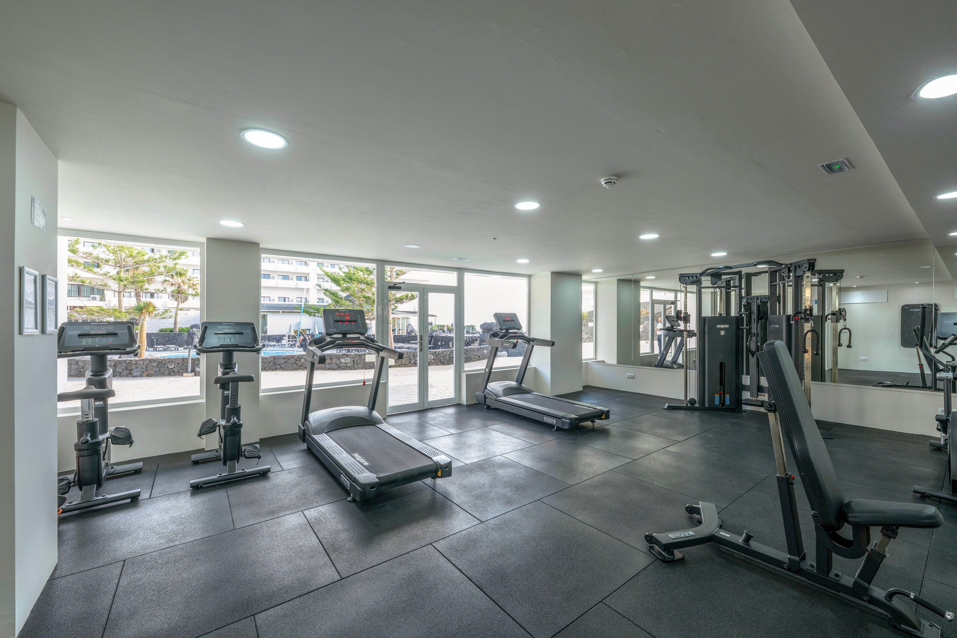 A spacious gym features several treadmills, stationary bikes, and strength training machines. The room is well-lit with large windows allowing natural daylight and views of the outdoors. The interior has a minimalist design with black flooring and white walls, creating a clean and professional atmosphere. No people are present, emphasizing the equipment and open space.
