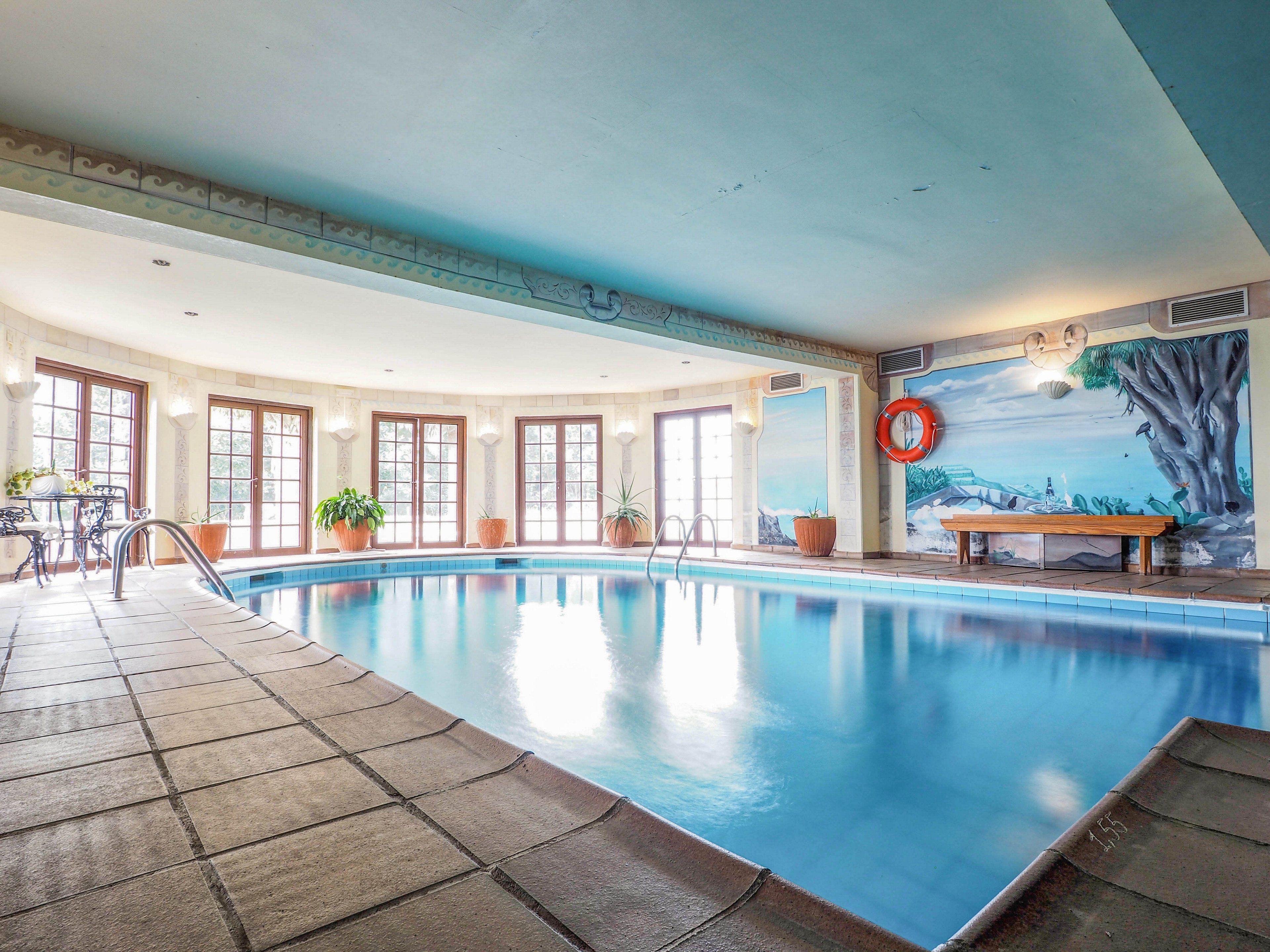A spacious indoor swimming pool is surrounded by tiled flooring and large windows letting in natural light. Potted plants and a small table with chairs add a touch of greenery and comfort to the setting. A painted wall mural depicting a scenic landscape and a mounted lifebuoy provide visual interest along one side of the room. The calm blue water and soft lighting create a relaxing, resort-like atmosphere.