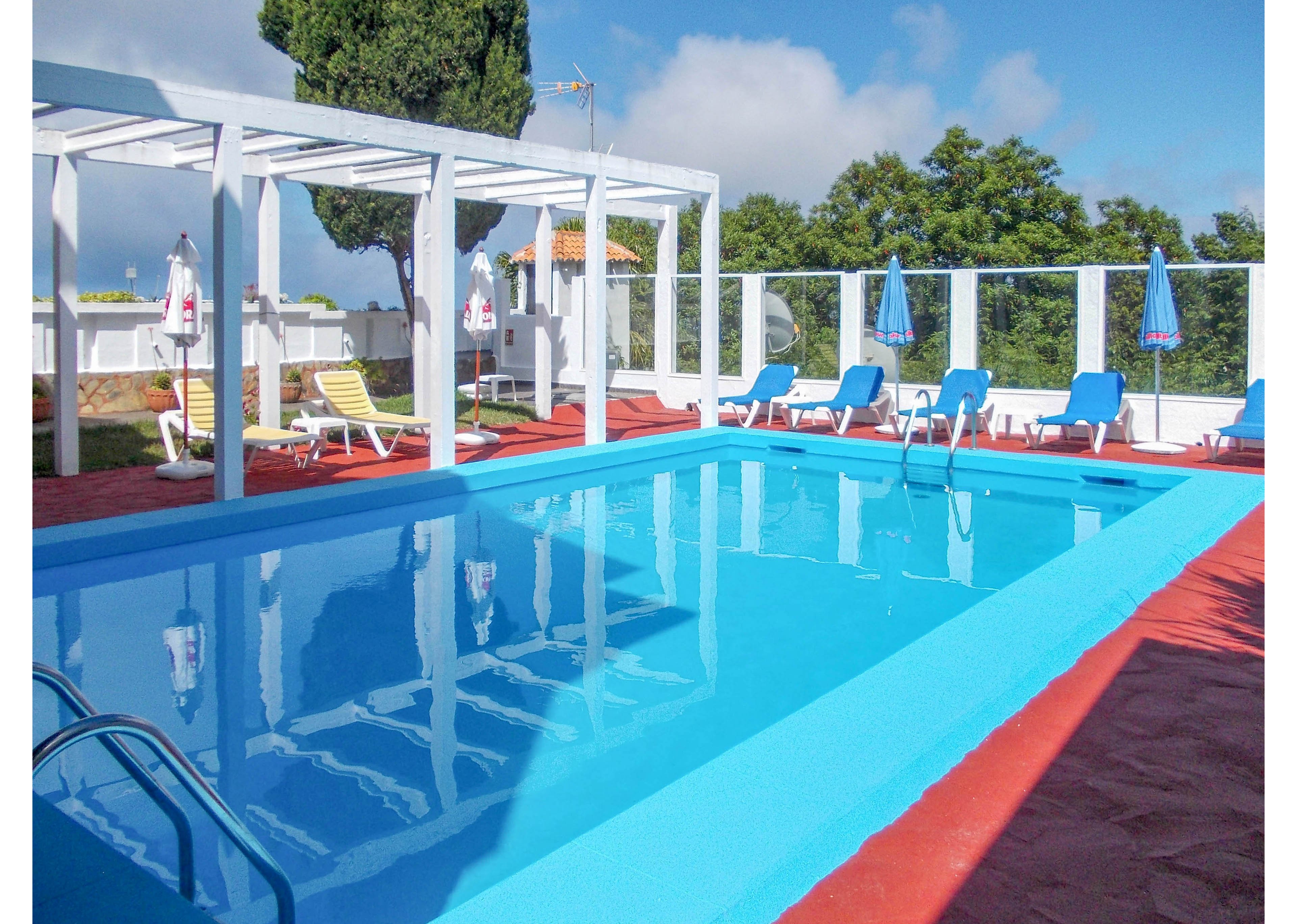 An outdoor swimming pool reflects the bright blue sky on a sunny day. Blue sun loungers and umbrellas are arranged around the pool on a red tiled deck. A white pergola structure and glass fence frame the pool area, with trees and a building roof visible in the background. The scene suggests a relaxing vacation or leisure setting in warm weather.