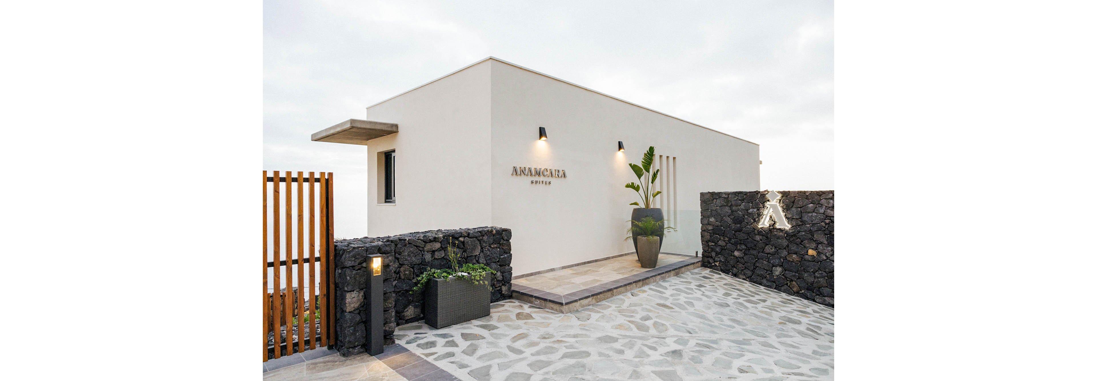 A contemporary hotel building features clean white walls and minimalist architecture. The entrance area is paved with stone tiles and decorated with large potted plants and dark stone walls. The name 'ANAKASTA SUITES' is clearly visible on the facade, along with a stylized 'A' sign illuminated on the stone wall. The setting appears serene and upscale, with soft outdoor lighting and a neutral color palette.