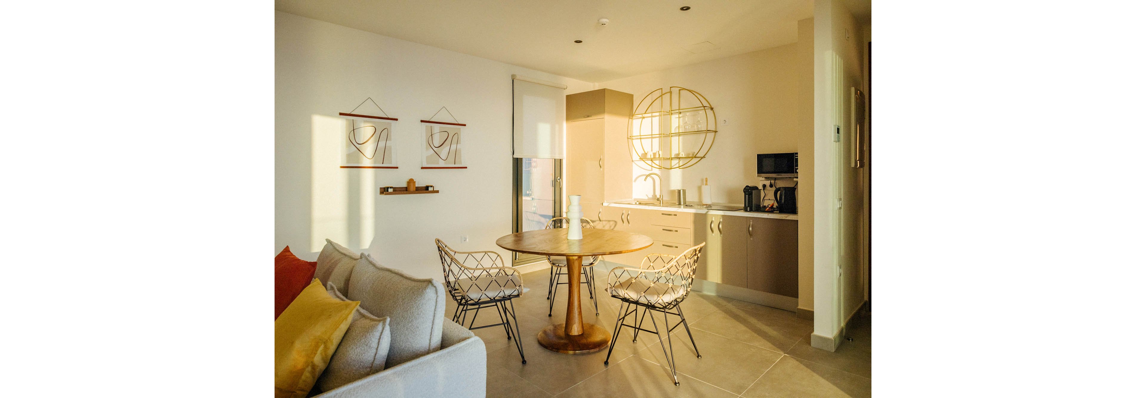 A contemporary kitchen and dining space is bathed in warm sunlight, featuring a round wooden table with metal chairs and minimalist wall art. The kitchen includes neutral cabinetry, a coffee maker, and open shelving with geometric design. Soft furnishings and muted tones create a cozy, inviting atmosphere.