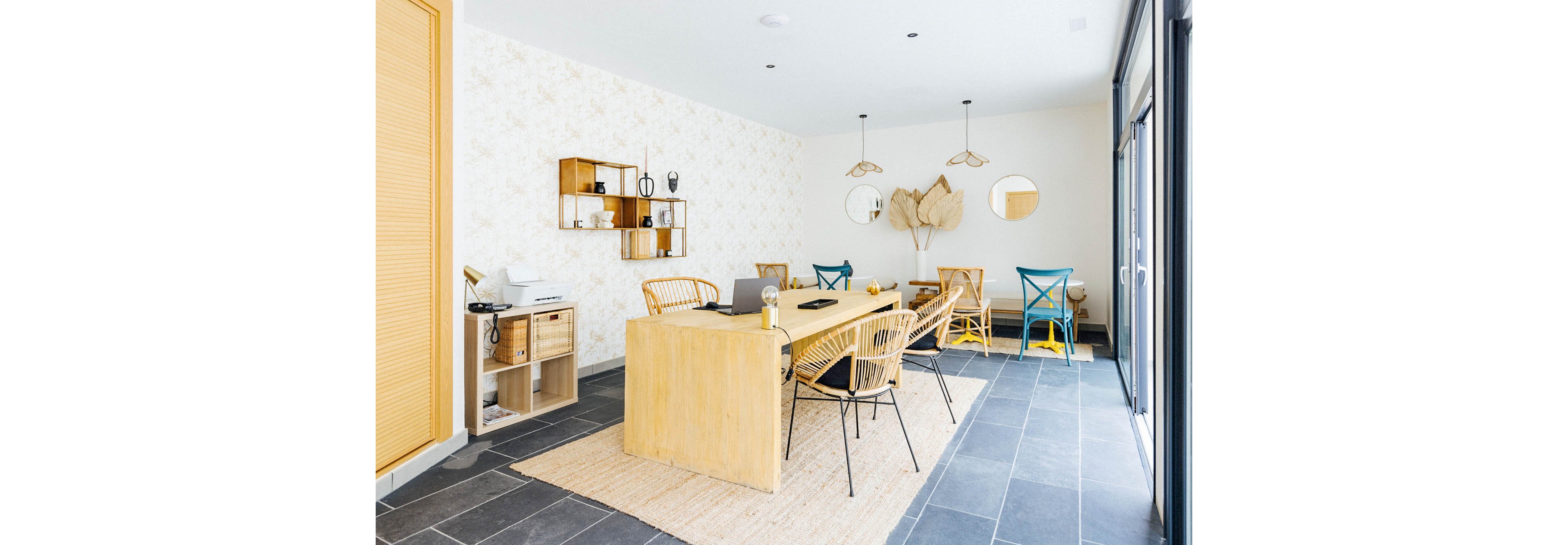 A bright, contemporary dining room features a large wooden table surrounded by wicker chairs. The space is decorated with minimalist shelving, neutral wallpaper, and pendant lighting. Natural light streams in through large windows, highlighting the black tile floor and woven rug. Decorative accents include dried leaves, mirrors, and ceramics, creating a calm and inviting atmosphere.