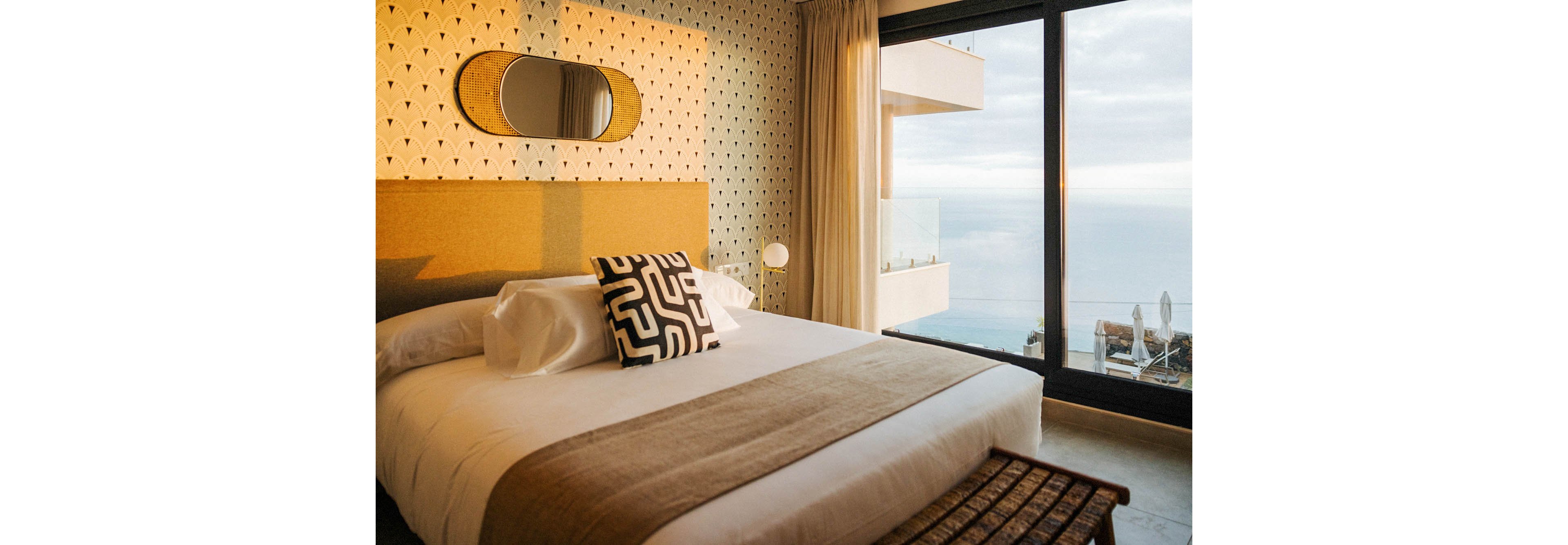 A contemporary hotel bedroom features a large bed with white linens and a bold geometric pillow. The room is decorated with patterned wallpaper, a stylish mirror, and neutral tones, creating a calm and inviting atmosphere. Floor-to-ceiling windows reveal a balcony and a scenic ocean view, enhancing the tranquil mood. The space is bathed in natural light, highlighting the modern furnishings and minimalist style.