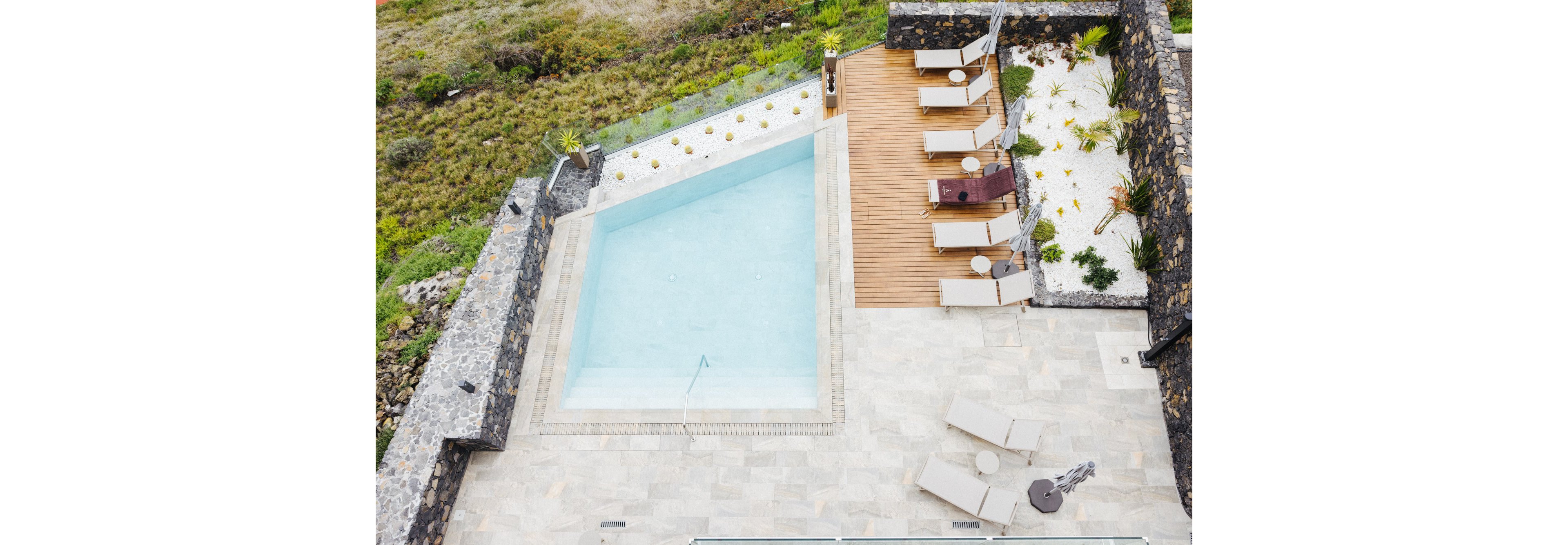 A geometric outdoor swimming pool is surrounded by a stone patio and wooden deck, featuring several beige sun loungers and small round tables. The setting is minimalist and modern, with landscaped rock gardens and greenery along the edges. The area appears tranquil and inviting, ideal for relaxation or sunbathing.