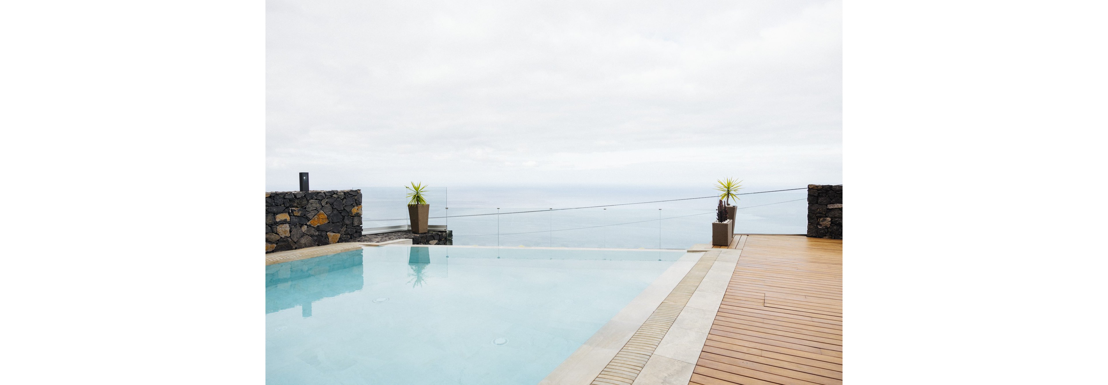 A serene infinity pool sits adjacent to a wooden deck, offering an unobstructed view of the ocean under a cloudy sky. The setting features minimalist stone planters with small plants and a glass safety barrier. The tranquil scene emphasizes relaxation and luxury in a contemporary outdoor space.
