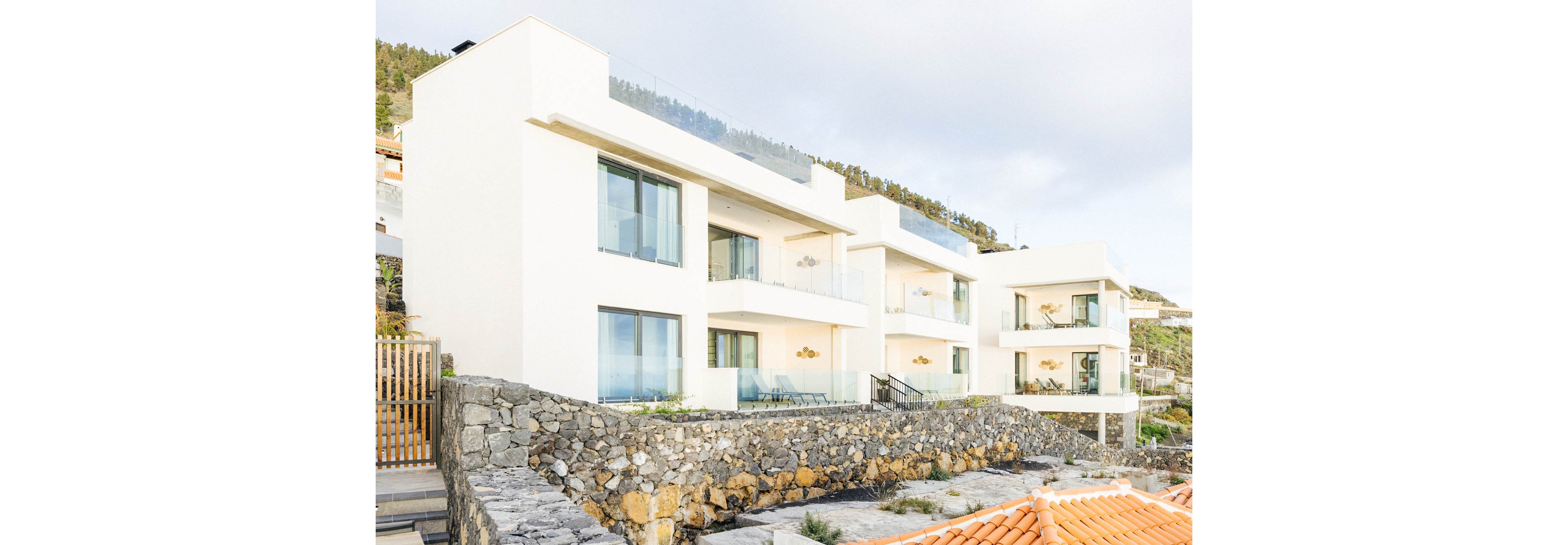 The image features a row of contemporary white apartment buildings with large glass balconies and expansive windows. The structures are set on a stone terrace with a scenic hillside in the background. The overall mood is bright and airy, with clean architectural lines and a minimalist aesthetic. No visible text, numbers, or identifiable brands are present in the image.