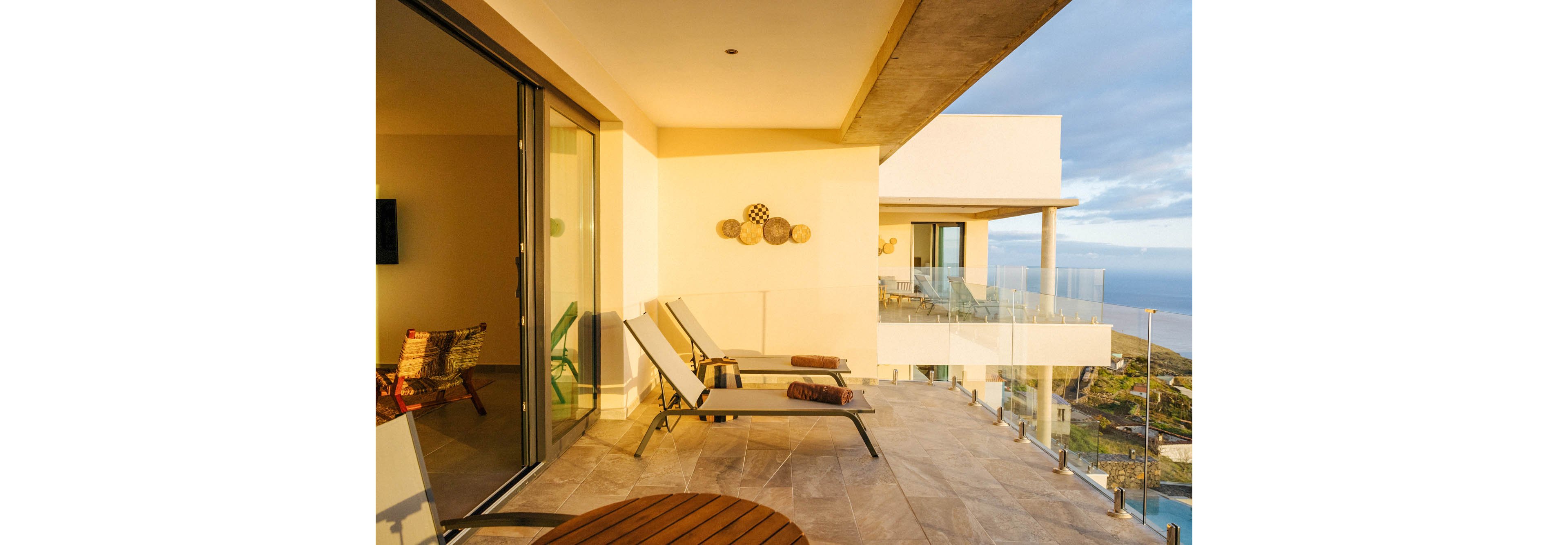 A spacious balcony features several reclining lounge chairs and a small round table, bathed in warm sunlight. The setting overlooks a scenic ocean view, with glass railings enhancing the open, airy feel. The decor includes woven wall art and neutral tones, creating a relaxed, contemporary atmosphere. Reflections in the glass doors reveal additional seating inside.