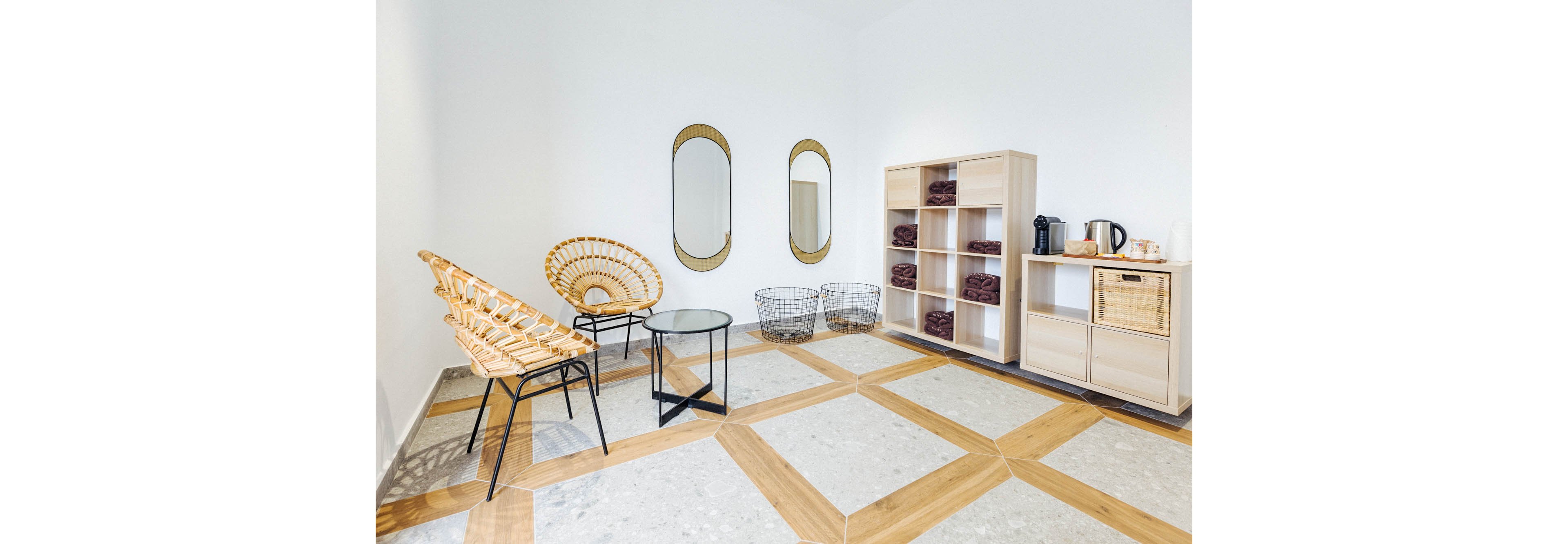A bright, minimalist waiting area features two rattan chairs and a glass-topped table set against a white wall. Three oval mirrors are mounted above two wire baskets, while a shelving unit holds neatly folded towels and a coffee station with a kettle. The geometric patterned floor and neutral color palette create a calm, modern atmosphere.
