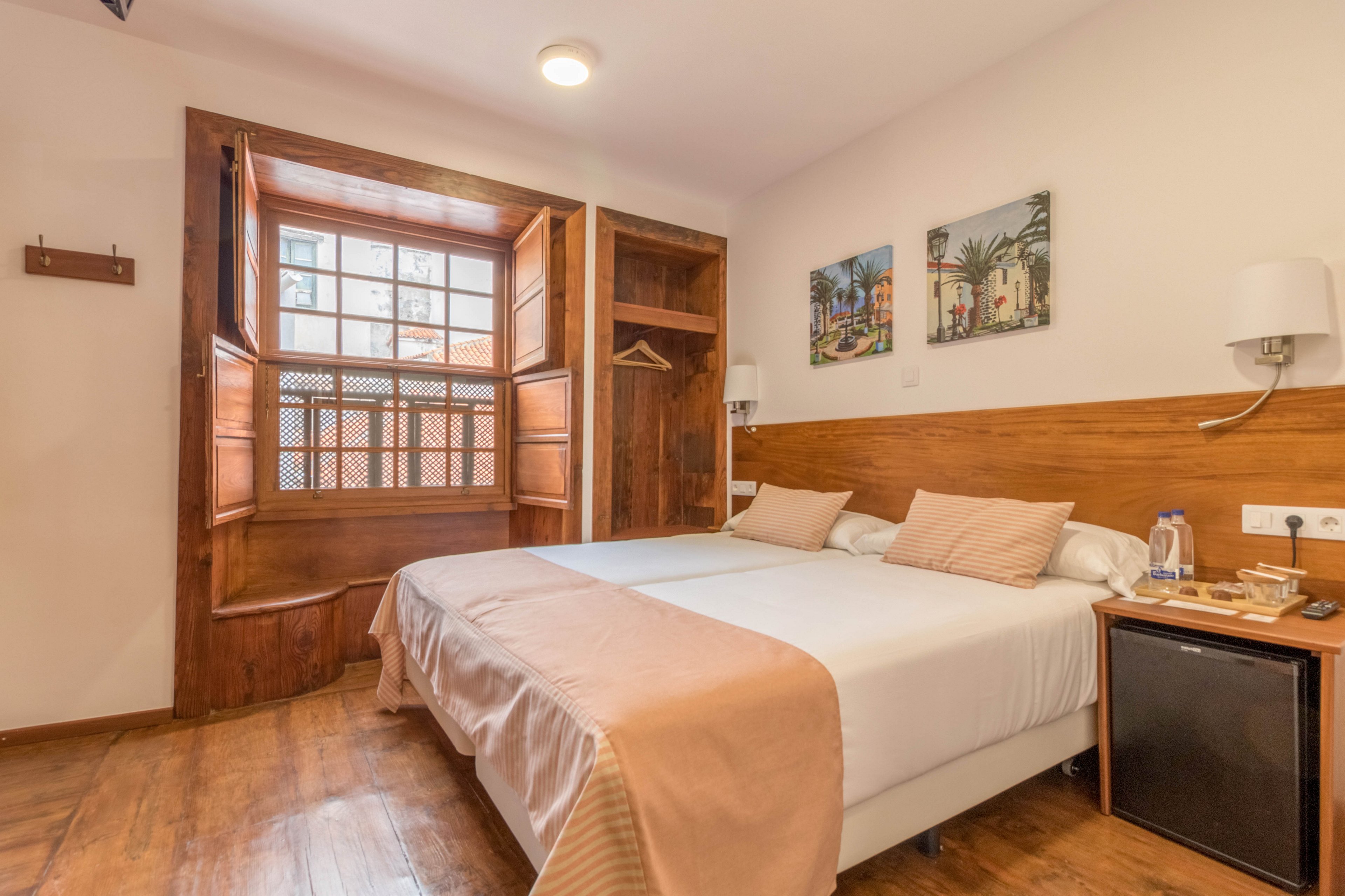 A bright hotel room features two twin beds pushed together, each with beige bedding and pillows. The space is accented by wooden floors, a large window with wooden shutters, and wall-mounted lamps. Artwork decorates the wall above the beds, and a small minibar with a kettle and cups sits beside the bed. The overall mood is warm and inviting, with natural light enhancing the cozy atmosphere.