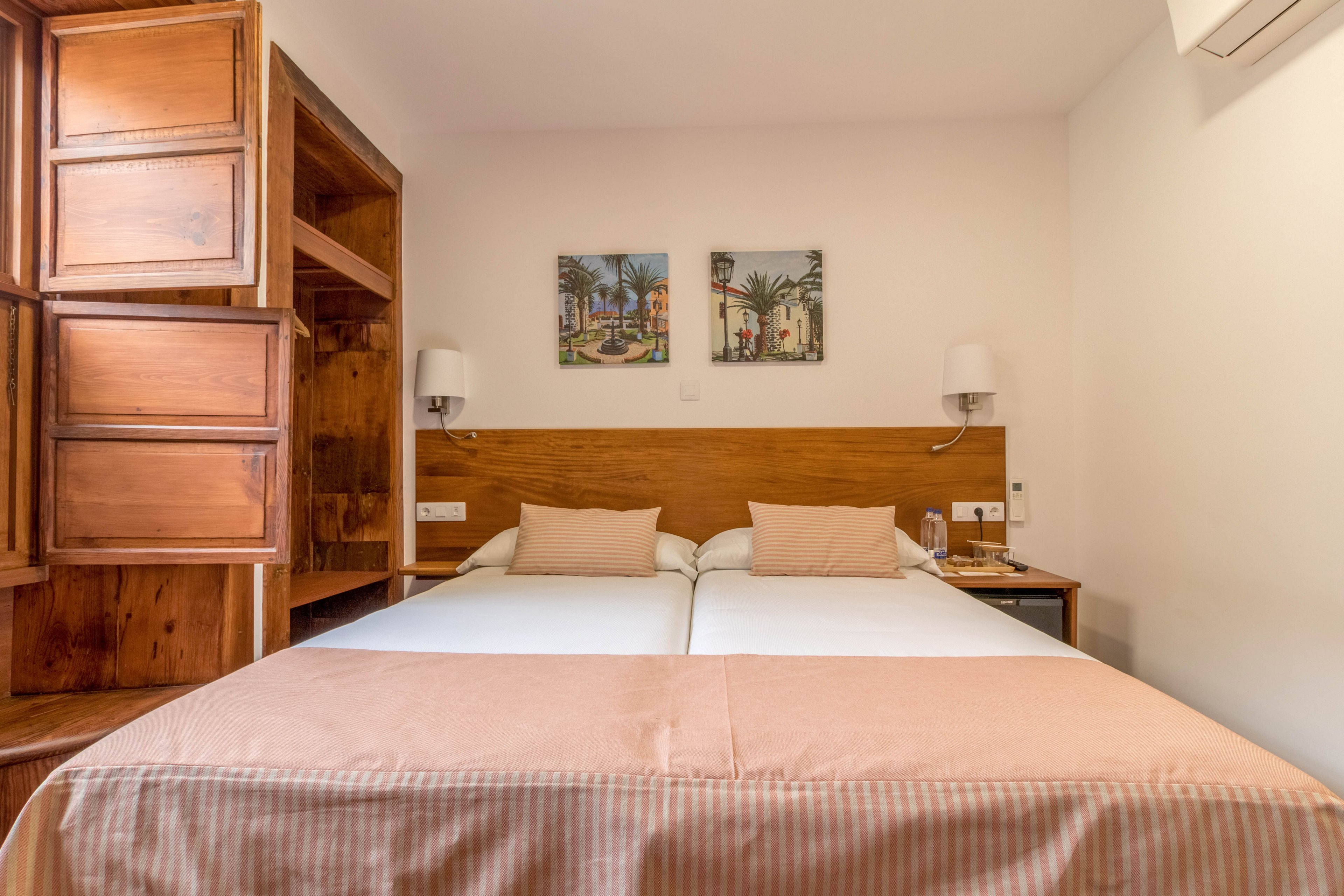 A neatly arranged hotel room features two twin beds pushed together, each with beige pillows and a matching bedspread. The room includes wooden furniture, such as an open wardrobe and bedside tables, and is decorated with two framed paintings above the headboard. Soft lighting from wall-mounted lamps and a neutral color palette create a calm, inviting atmosphere. An air conditioning unit is visible on the upper right wall.