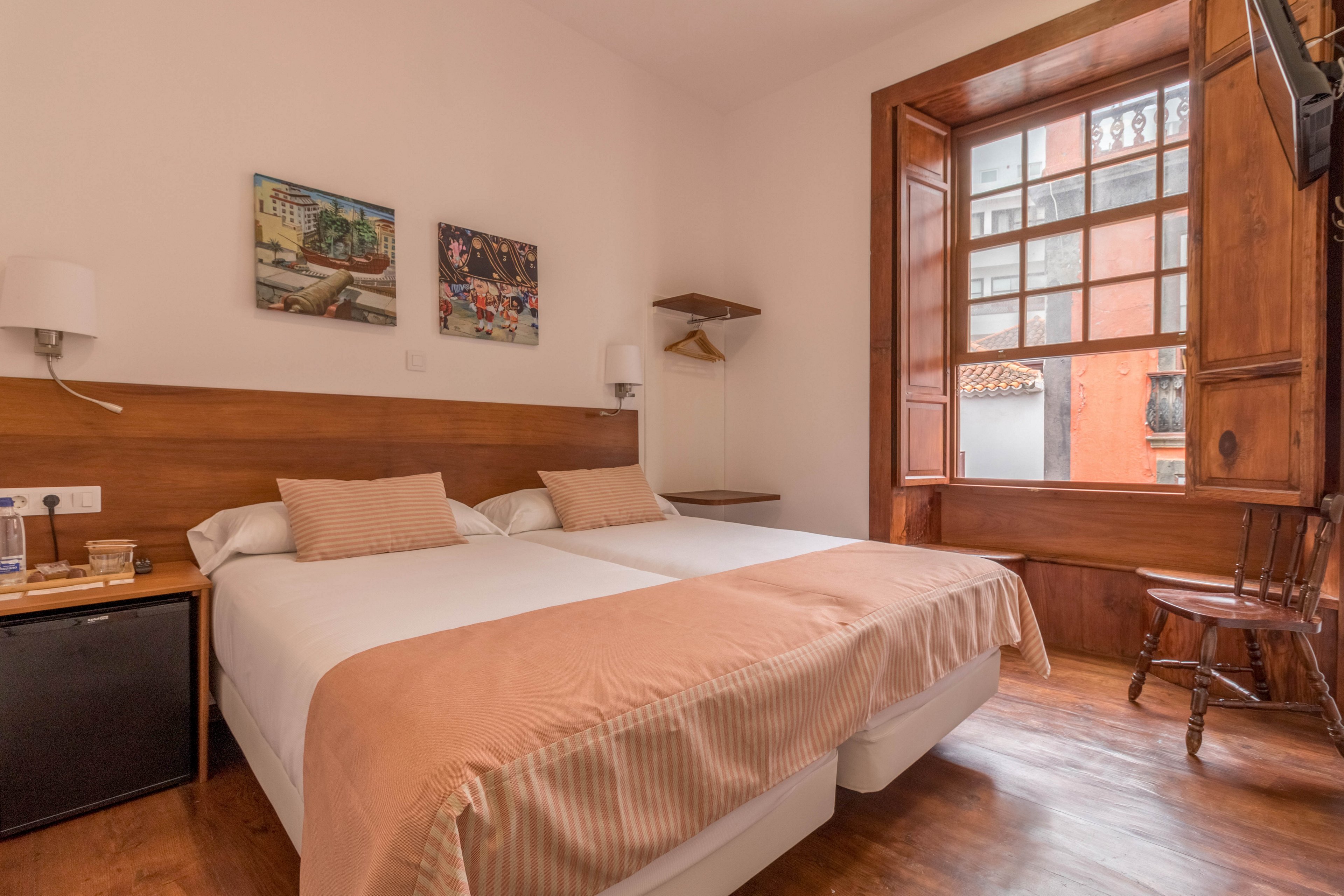 A cozy hotel room features two twin beds with beige throws and pillows, set against a wooden headboard. The space is illuminated by natural light from a large window with wooden framing, offering a view of an adjacent building. The room includes a small nightstand, a mini fridge, wall-mounted lamps, and two paintings above the beds. The overall atmosphere is warm and inviting, with neutral tones and simple decor.