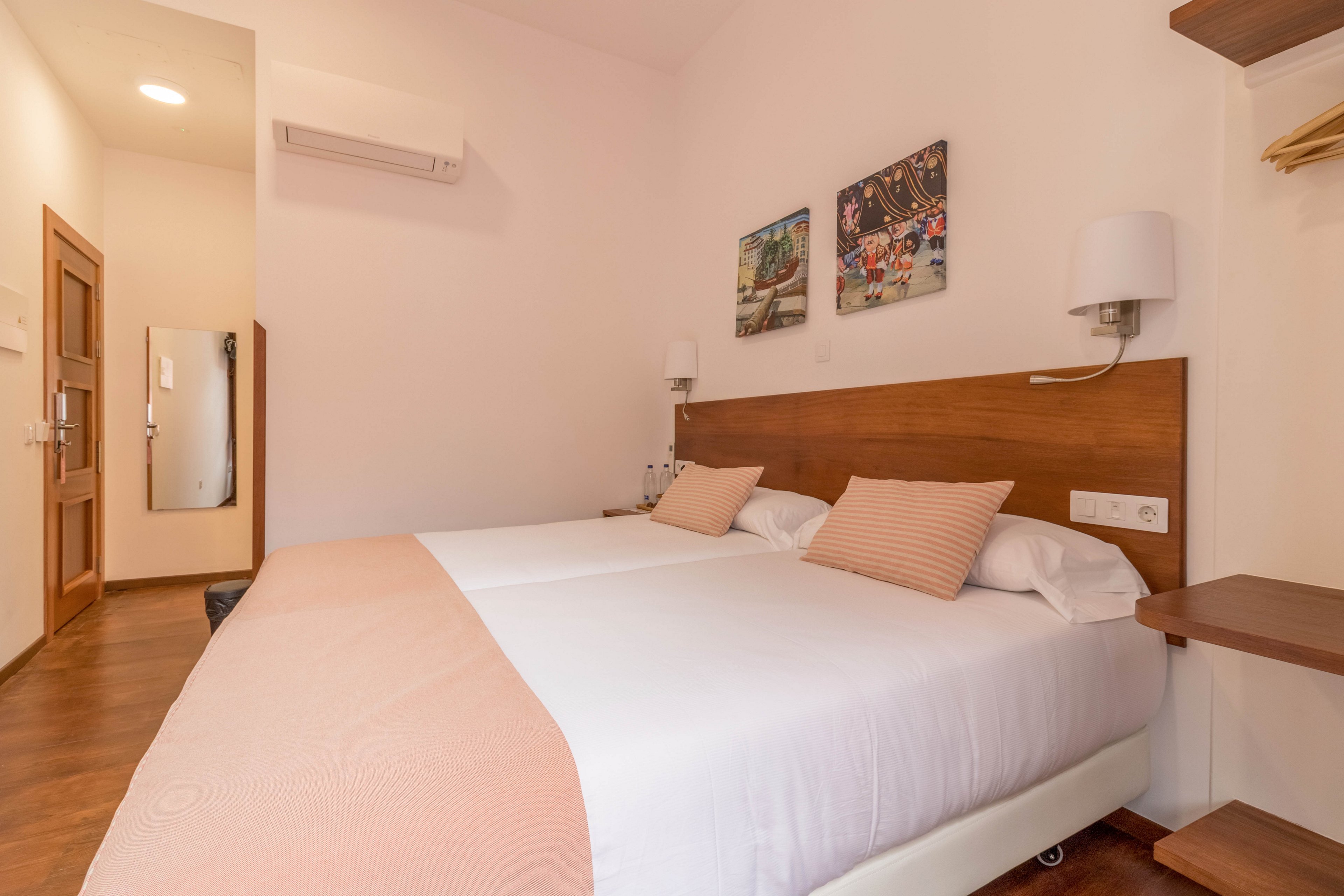 A clean, modern hotel room features two twin beds with white linens and soft peach accents. The space is decorated with contemporary wall art and equipped with bedside lamps and shelves. A wall-mounted air conditioner and a large mirror are visible, enhancing the room's functionality. The overall palette is light and inviting, creating a comfortable atmosphere for guests.
