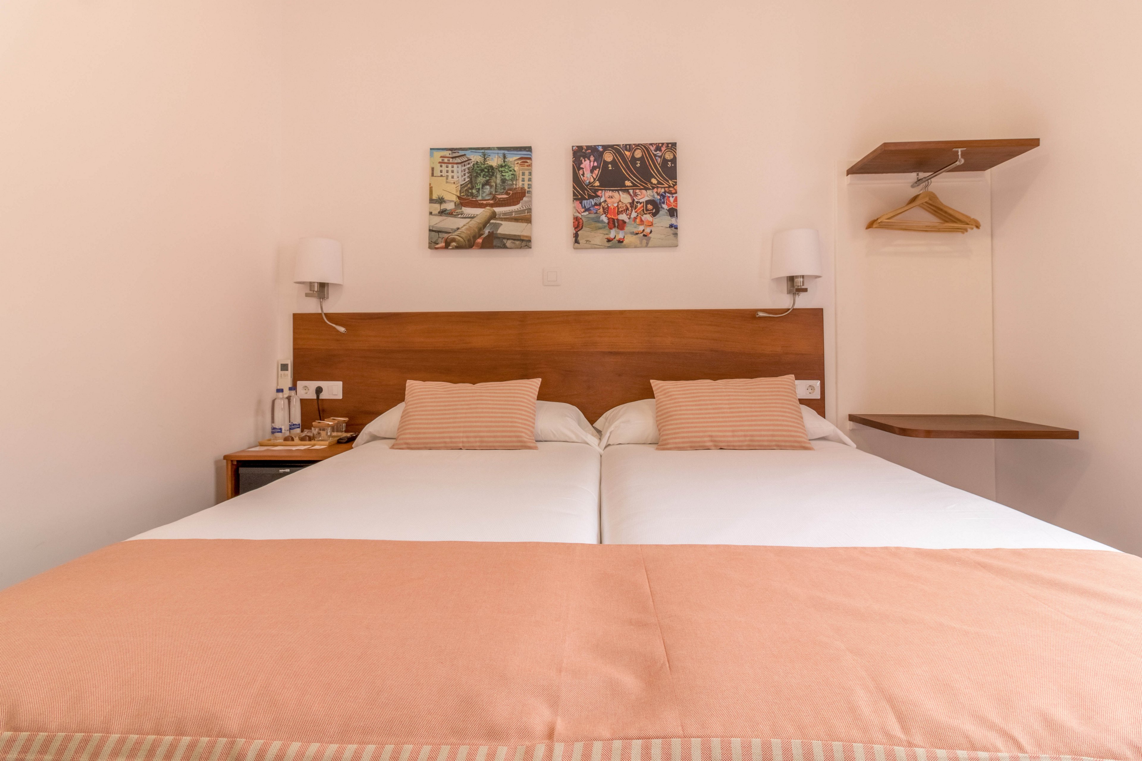 A neatly arranged hotel room features two twin beds with peach-colored bedding and matching pillows. The setting is minimalistic, with a wooden headboard, wall-mounted lamps, and two framed artworks above the beds. A small side table holds water bottles and glasses, while an open shelf and hanger are visible on the wall. The overall palette is warm and inviting, creating a comfortable and relaxing atmosphere.