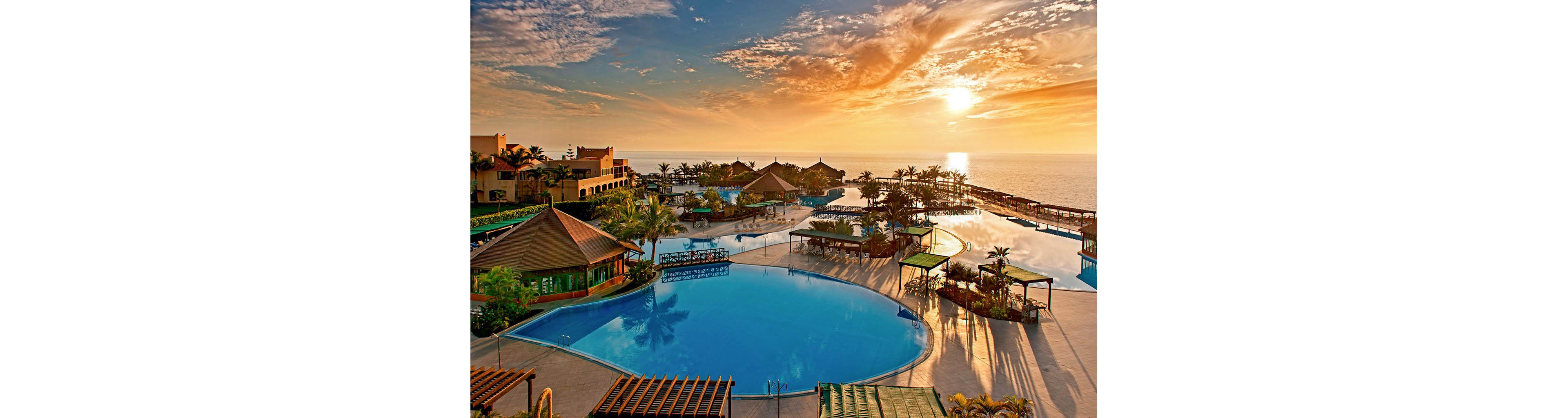 A large seaside resort features expansive blue swimming pools overlooking the ocean at sunset. The scene includes multiple pool levels, palm trees, and open-air structures arranged along the waterfront. Warm golden light reflects off the calm water and highlights the surrounding terraces. The sky is filled with dramatic clouds as the sun nears the horizon, creating a tranquil vacation atmosphere.