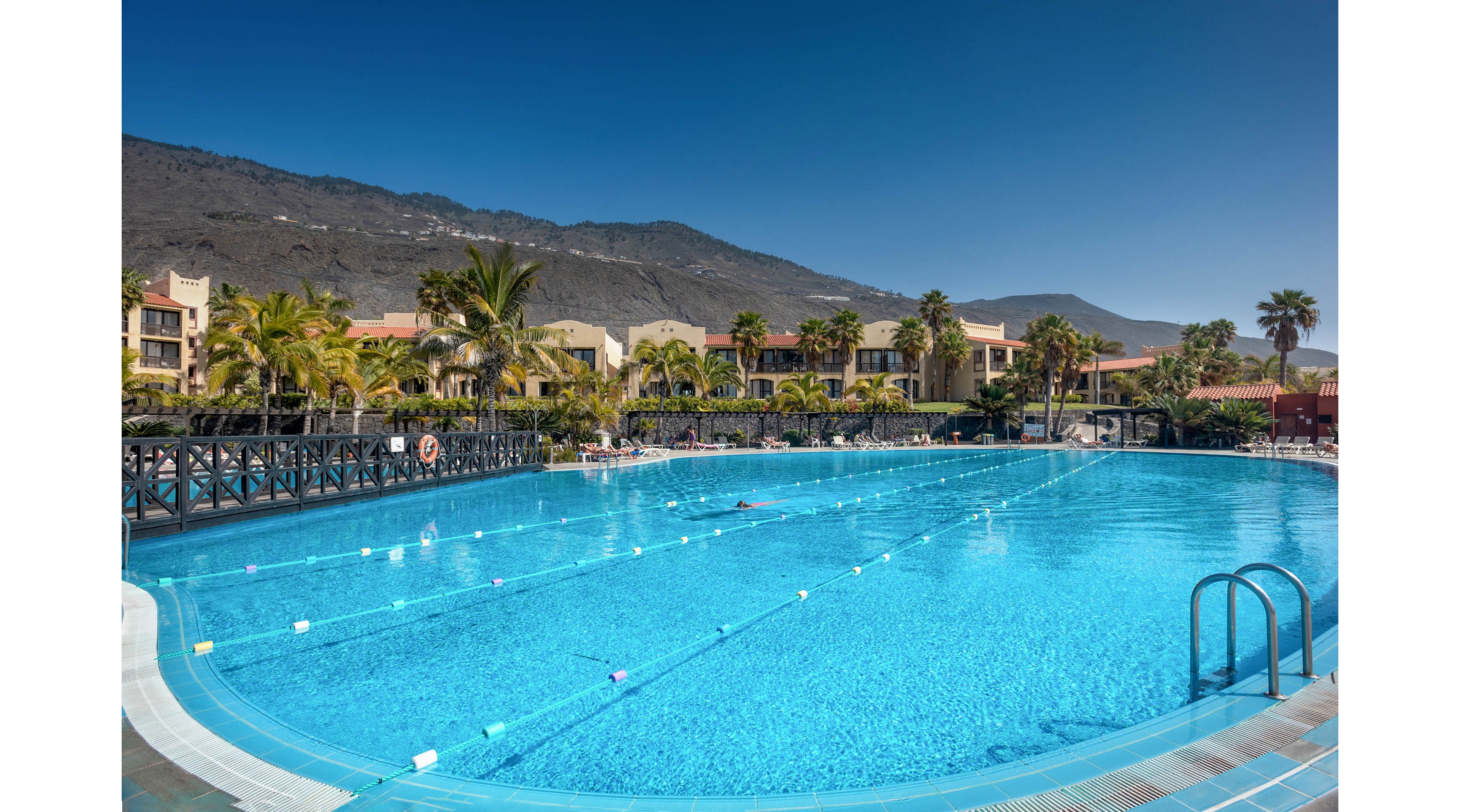 A large outdoor swimming pool glistens under a clear blue sky at a resort. Low-rise hotel buildings and palm trees surround the pool, creating a tropical atmosphere. Floating lane markers stretch across the water, and metal ladders provide access at the pool edge. In the background, dark mountains rise above the resort, contrasting with the bright turquoise water.