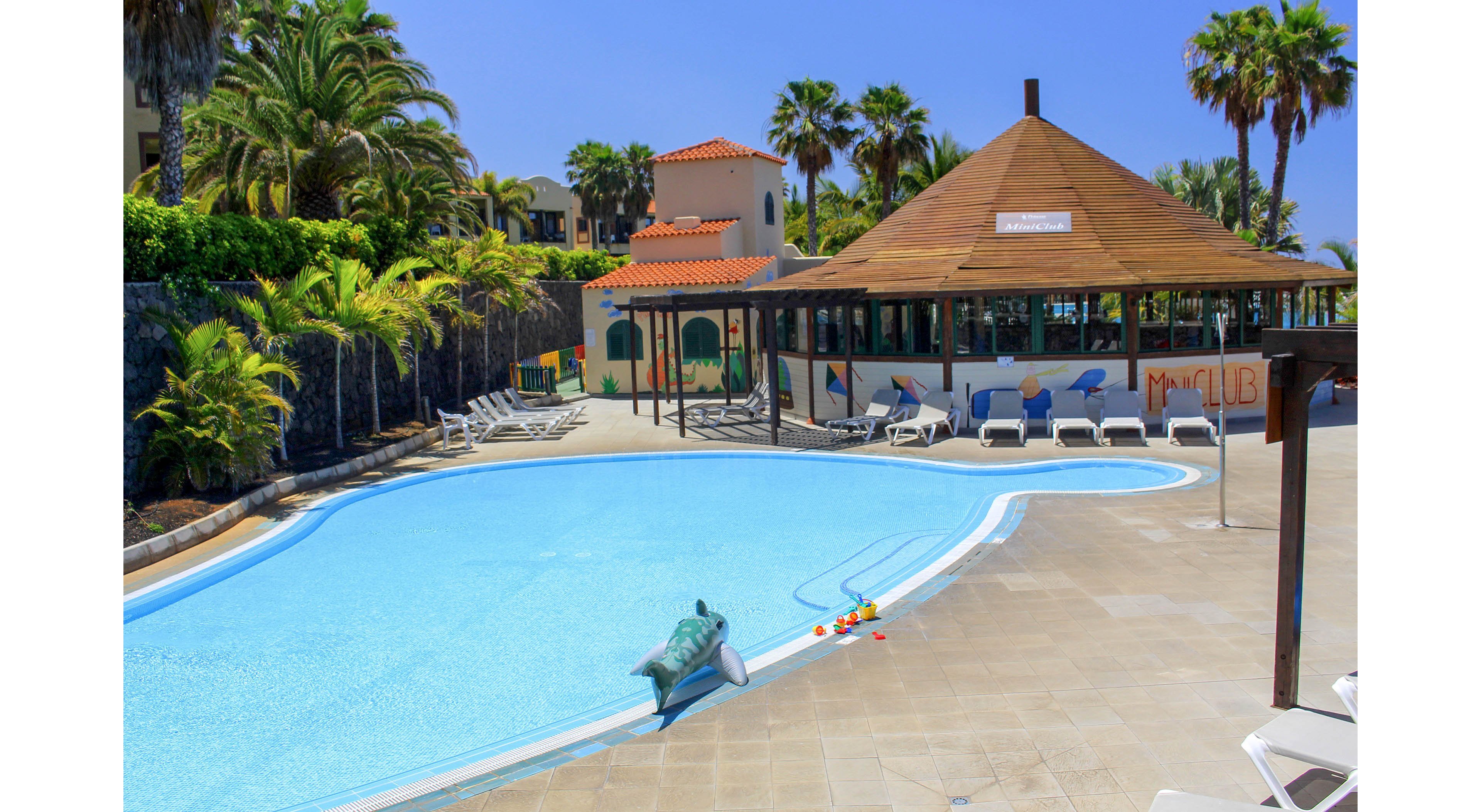 An outdoor resort swimming pool is shown under bright sunshine, surrounded by palm trees and lush greenery. A wooden-roofed poolside bar and adjacent building with murals stand beside the water, with several white lounge chairs arranged around the deck. A small dolphin-shaped fountain or sculpture is placed near the pool edge, adding a playful touch. The scene suggests a relaxing vacation atmosphere in a warm, tropical setting.
