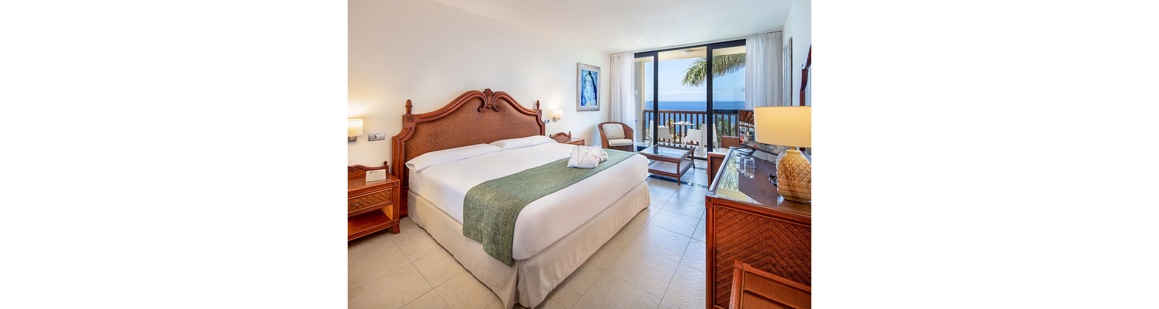A spacious hotel room features a large bed with white linens and a green runner, accented by carved wooden furniture. The room opens onto a balcony with a view of the ocean and palm trees through large glass doors. Soft lighting from bedside lamps and a table lamp creates a warm, inviting atmosphere. Neutral walls, tiled flooring, and simple decor contribute to a clean, relaxing coastal style.