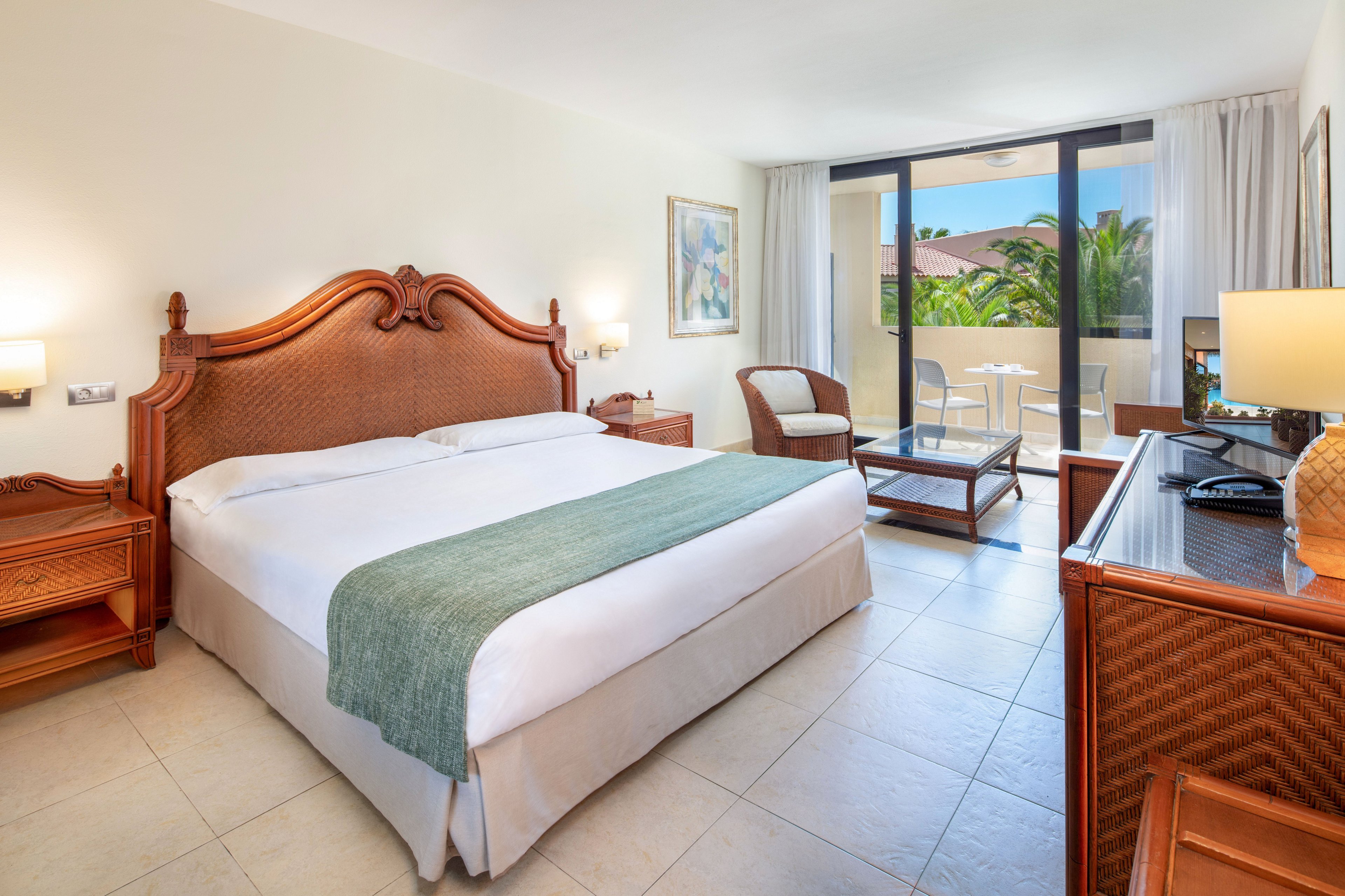 Spacious hotel room featuring a large double bed with carved wooden headboard and a light green throw. The setting includes tiled floors, wicker-style furniture, and a desk with lamps, creating a warm and inviting atmosphere. Large glass doors open to a balcony with outdoor seating and views of lush greenery and palm trees. Natural daylight fills the room, highlighting its clean, comfortable, and relaxing style.