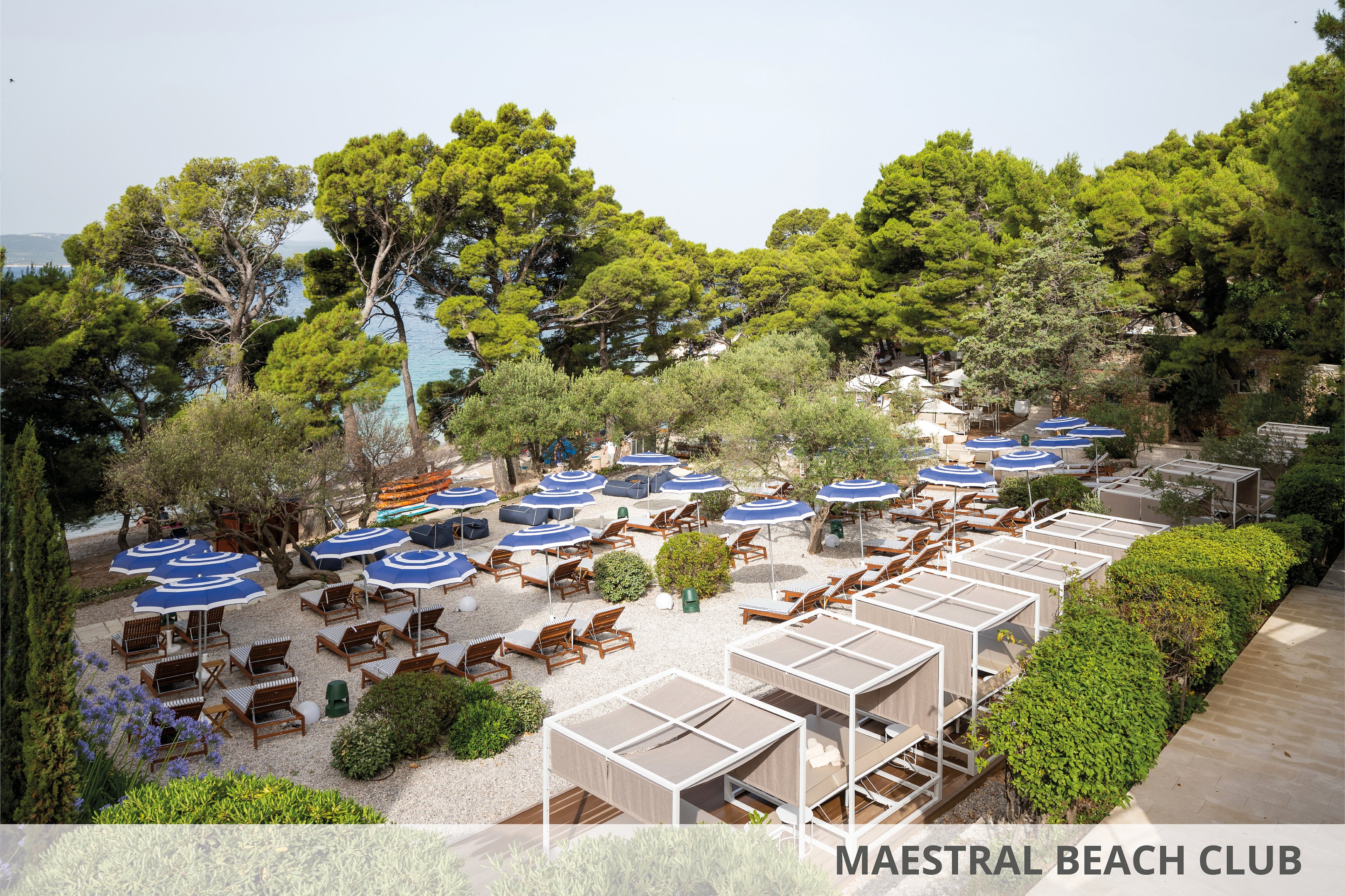 A serene outdoor setting featuring wooden lounge chairs and blue umbrellas arranged on a pebble beach. Cabanas with white frames are positioned alongside lush greenery, overlooking a coastal area with tall pine trees. The scene evokes relaxation and leisure in a natural environment.
