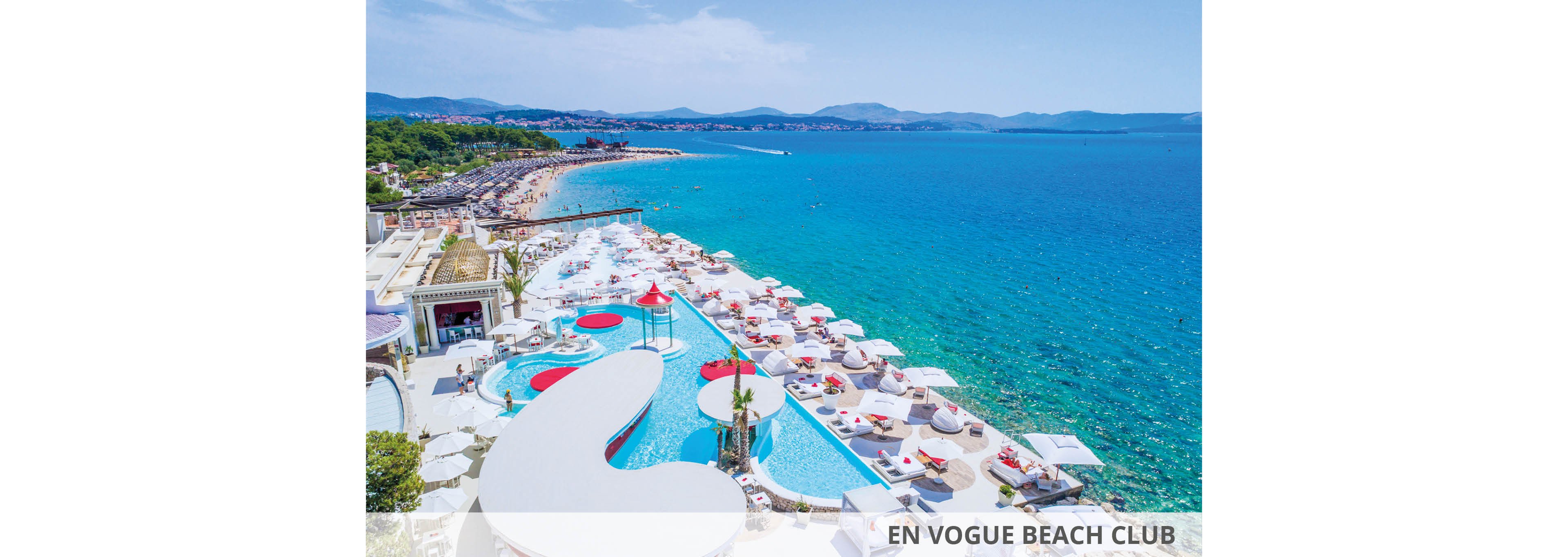 A vibrant beach club featuring multiple pools and white sun loungers overlooking a stunning turquoise ocean. The setting includes a spacious deck area with umbrellas and seating arrangements, ideal for relaxation and leisure. Visible text reads 'EN VOGUE BEACH CLUB,' emphasizing the upscale ambiance.