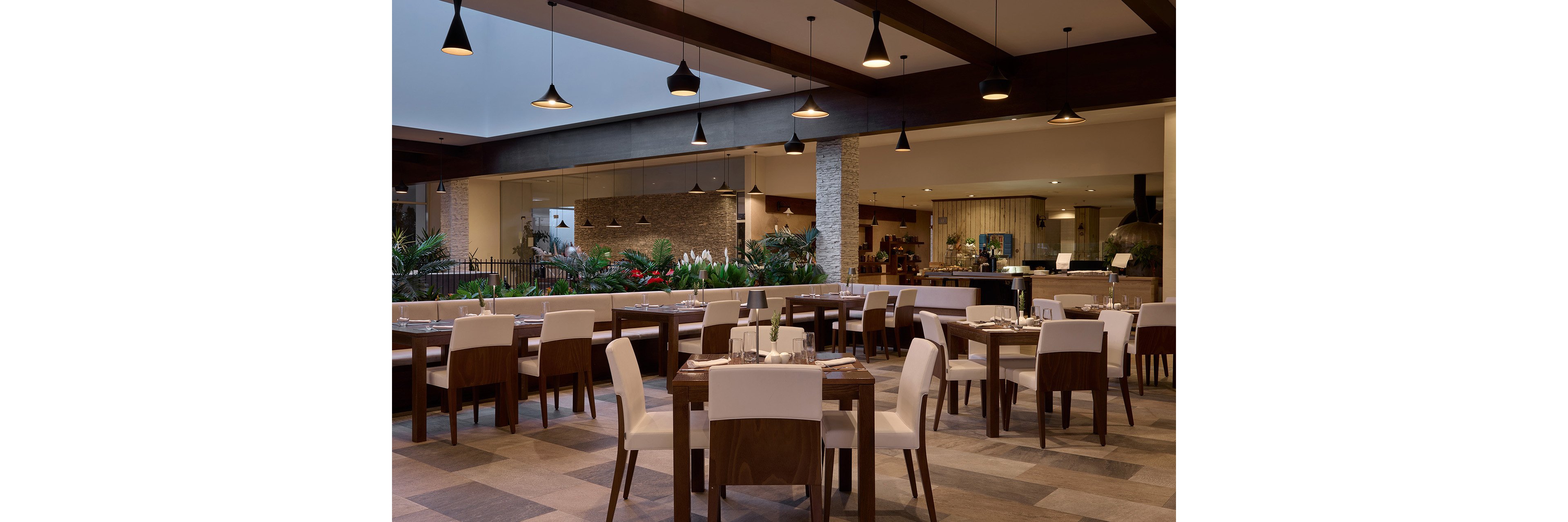 A contemporary restaurant dining area features neatly arranged wooden tables with white upholstered chairs. The space is open-concept, with a view into adjacent sections of the venue and a landscaped area with green plants. Warm pendant lighting hangs from dark wooden beams, creating a cozy yet upscale atmosphere. Neutral tones and clean lines emphasize a modern interior design style.