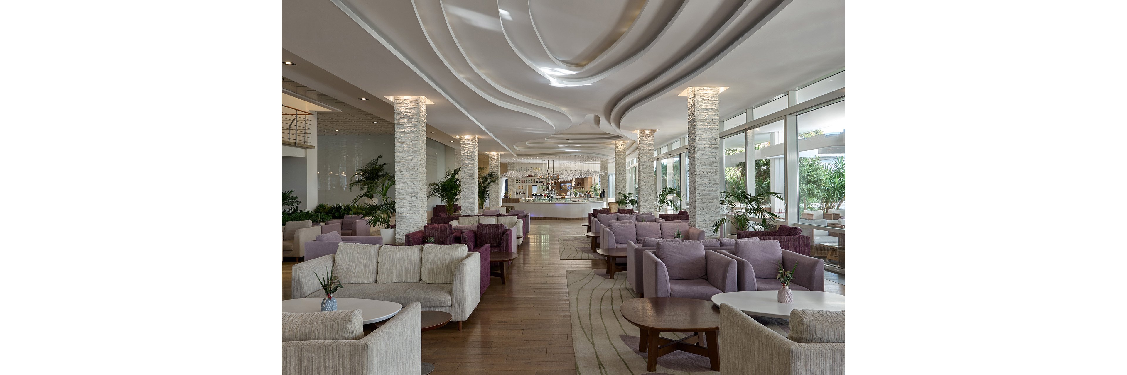 A spacious modern lounge features rows of cushioned armchairs and sofas arranged around low circular tables. The setting appears to be a hotel or resort lobby bar with a counter visible in the background. Large windows on one side let in natural light and reveal lush greenery outside. Decorative ceiling waves, stone-clad columns, and wooden flooring create a sophisticated, contemporary atmosphere.