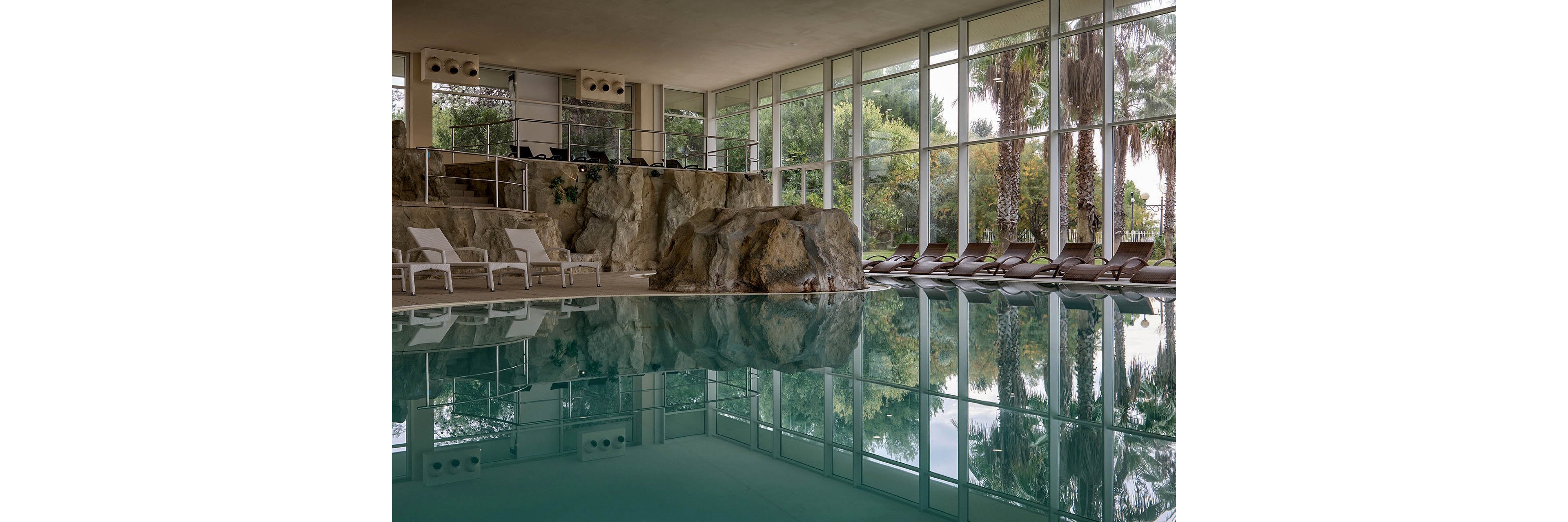 An indoor swimming pool reflects large floor-to-ceiling windows and lush greenery outside. A natural rock formation rises beside the pool, adding a spa-like, resort atmosphere. Rows of empty lounge chairs line the far side, suggesting a quiet, relaxing environment. Soft, neutral tones and calm water create a serene, tranquil mood.