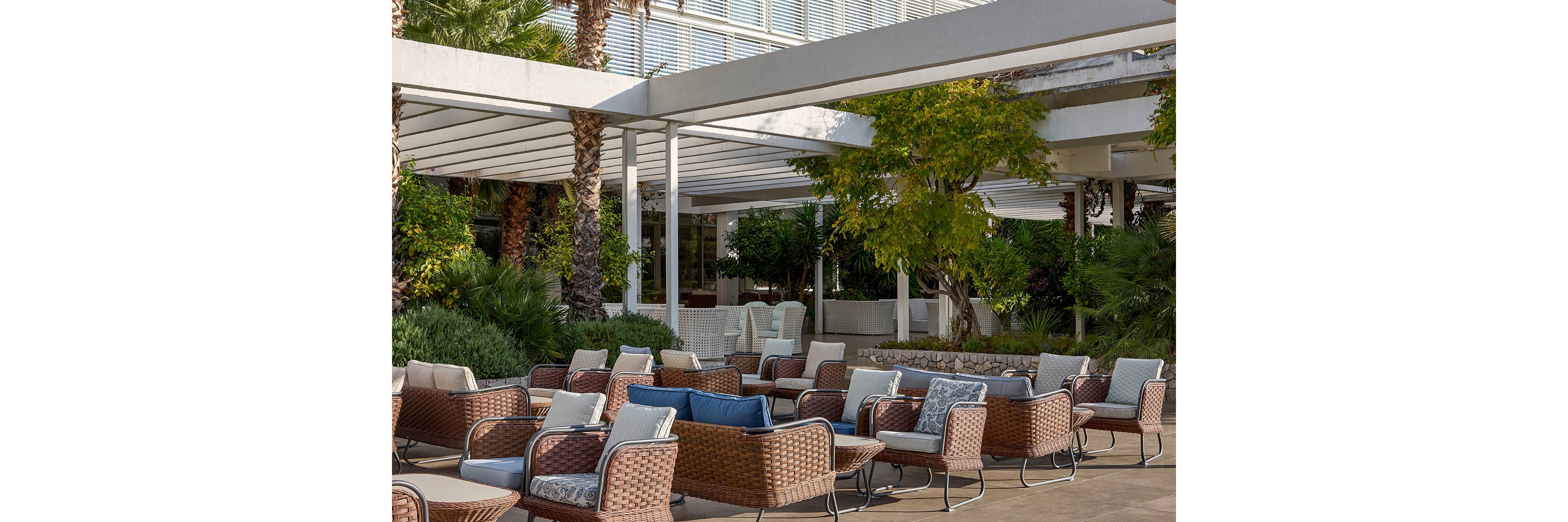 An outdoor lounge area features numerous wicker armchairs with cushioned seats arranged in clusters. The setting appears to be a modern patio or hotel terrace surrounded by lush greenery and palm trees. White pergola structures provide partial shade over the seating area, creating a relaxed and inviting atmosphere. Neutral and blue cushions add subtle color accents to the contemporary design.