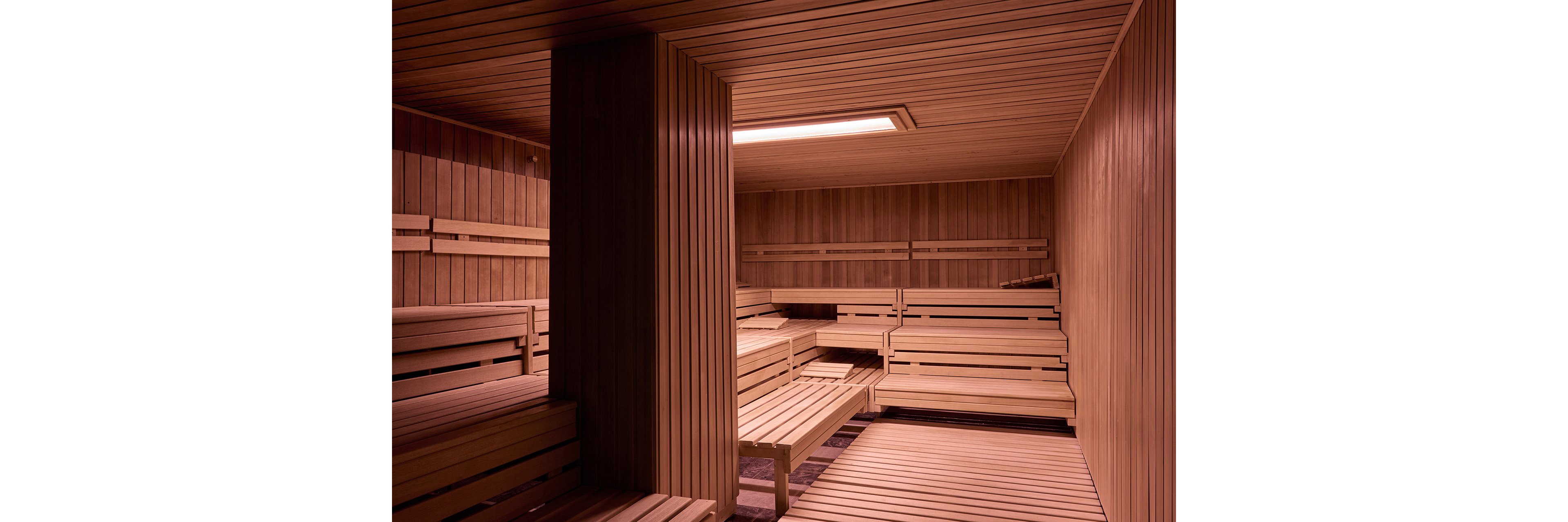 A spacious wooden sauna interior is shown completely empty. Multiple tiers of benches line the walls, creating ample seating on both sides of the room. Warm overhead lighting casts a soft glow on the smooth wooden panels and slats. The scene feels clean, orderly, and ready for relaxation in a spa or wellness setting.