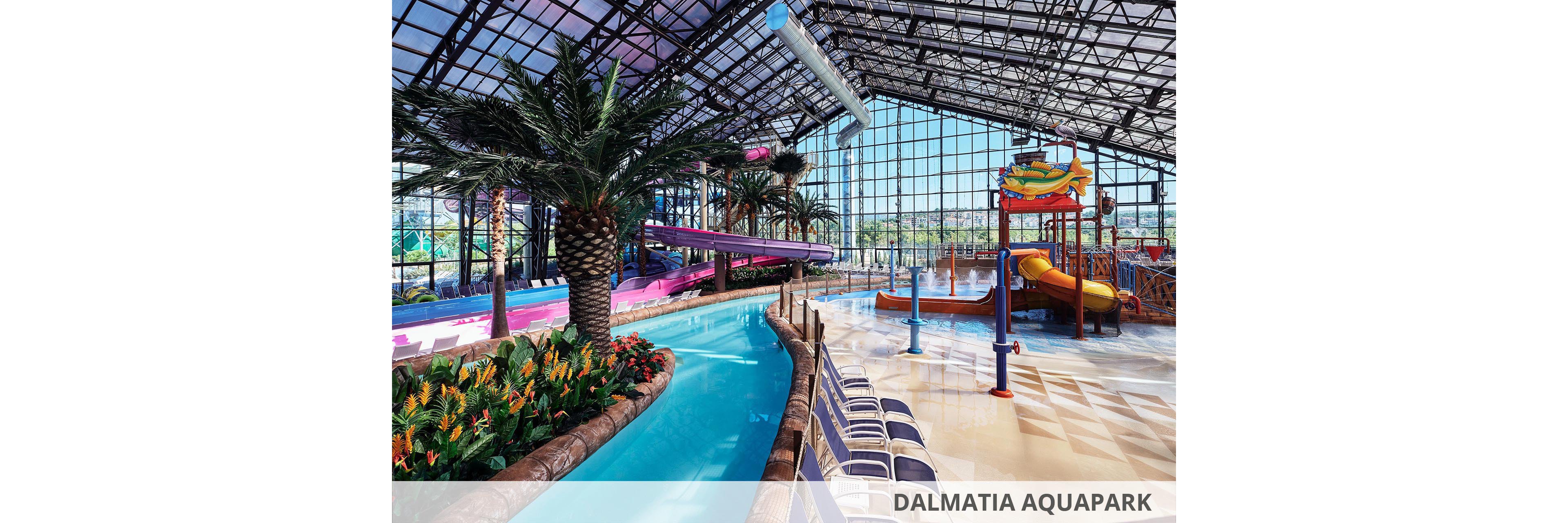 An expansive indoor water park features a winding lazy river bordered by lush tropical plants and tall palm trees. Colorful water slides and a multi-level play structure with fountains and sprayers stand in the background. Rows of empty lounge chairs line the poolside under a high glass ceiling that lets in abundant natural light. The scene feels bright, clean, and family-friendly, suggesting a resort or recreational attraction.