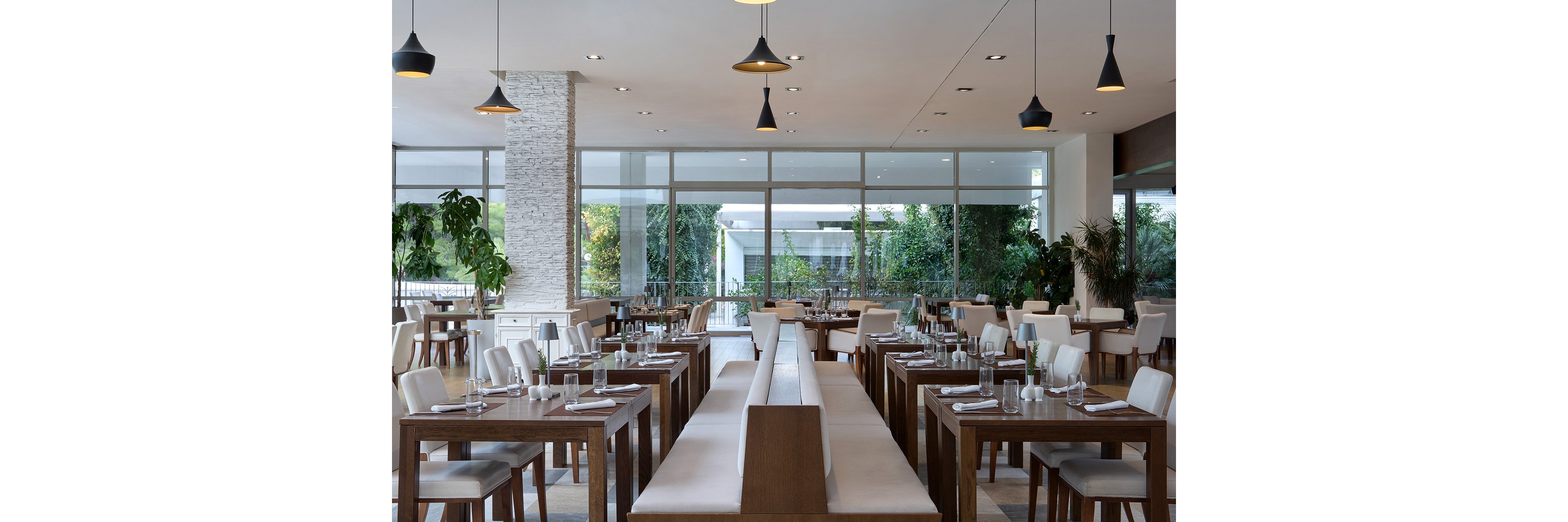 A contemporary restaurant dining area features rows of wooden tables and cushioned bench seating. Large floor-to-ceiling windows let in natural light and reveal lush greenery outside. Pendant lights hang from the ceiling, adding a modern touch to the clean, minimalist design. Tables are neatly set with glassware and napkins, creating a calm and inviting atmosphere.