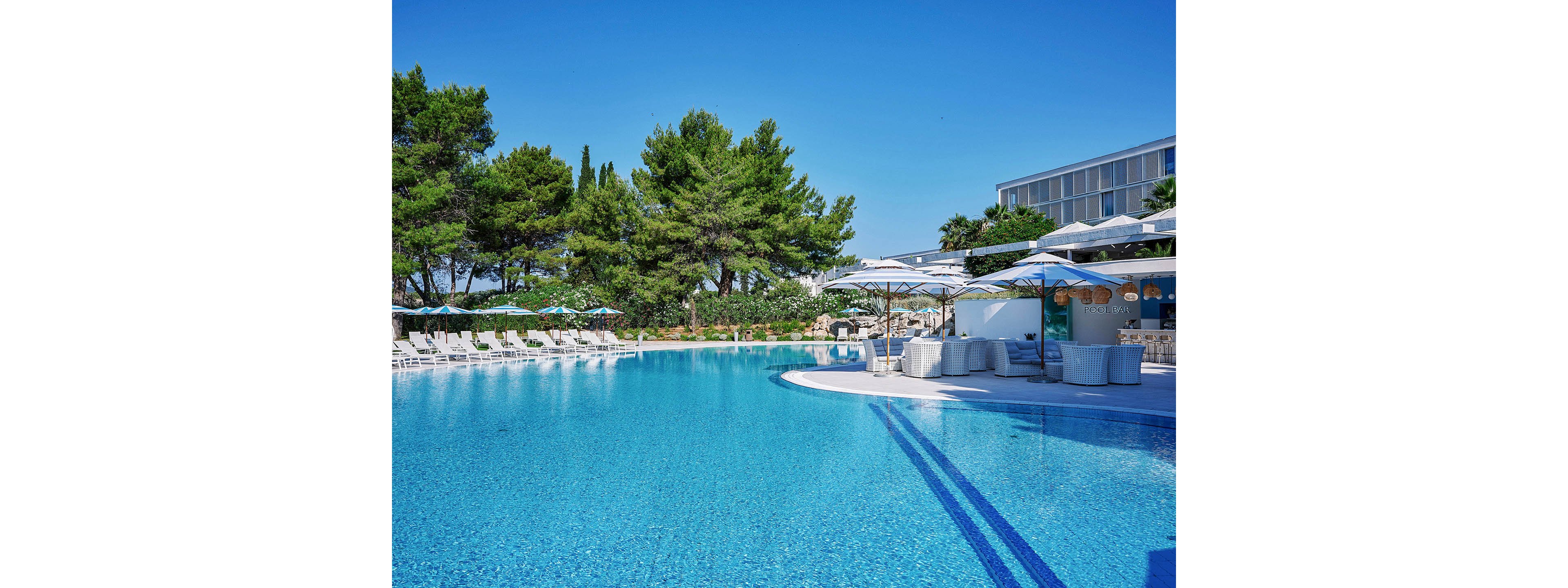 A large, clear blue swimming pool stretches across the foreground of a sunny resort. White lounge chairs and umbrellas line the poolside, creating a relaxing vacation atmosphere. Tall green trees and landscaped greenery form a natural backdrop behind the pool area. A modern building and shaded seating area with parasols are visible on the right, suggesting a hotel or resort complex.