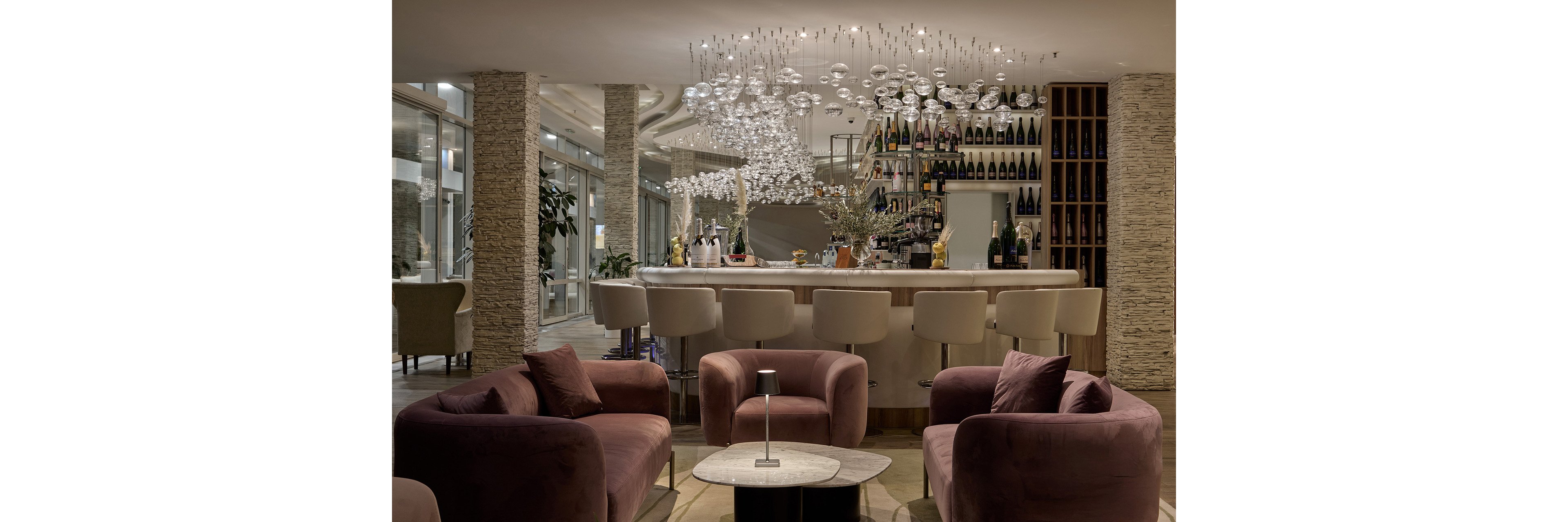A sophisticated hotel bar interior features a sleek white counter lined with cushioned bar stools. Plush mauve armchairs surround a small round table in the foreground, creating an inviting lounge area. The back bar is filled with neatly arranged bottles and glassware, while a sculptural chandelier of hanging glass orbs adds a dramatic focal point. Floor-to-ceiling windows and stone-textured columns contribute to the contemporary, upscale atmosphere.
