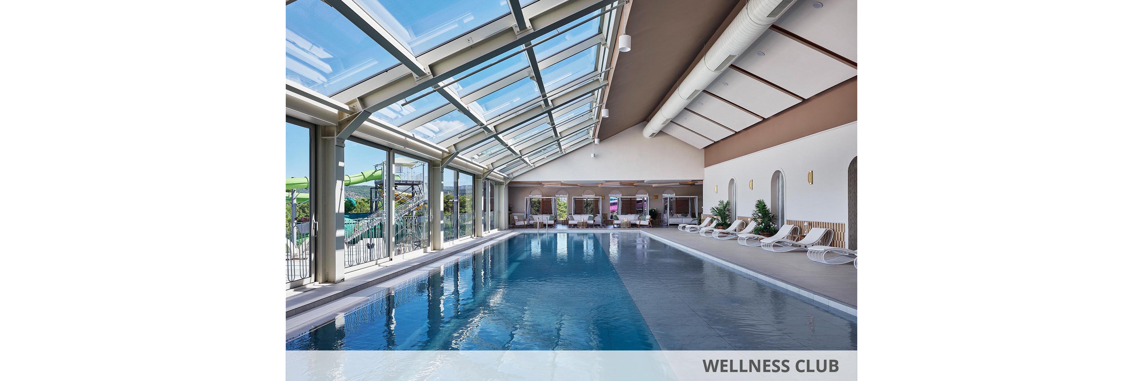 A spacious indoor swimming pool stretches along a bright, modern room. Large floor-to-ceiling windows and a glass roof let in abundant natural light and views of an adjacent water park. White lounge chairs line the far wall, creating a relaxing resort atmosphere. The clean architecture and blue water emphasize a calm, contemporary leisure setting.