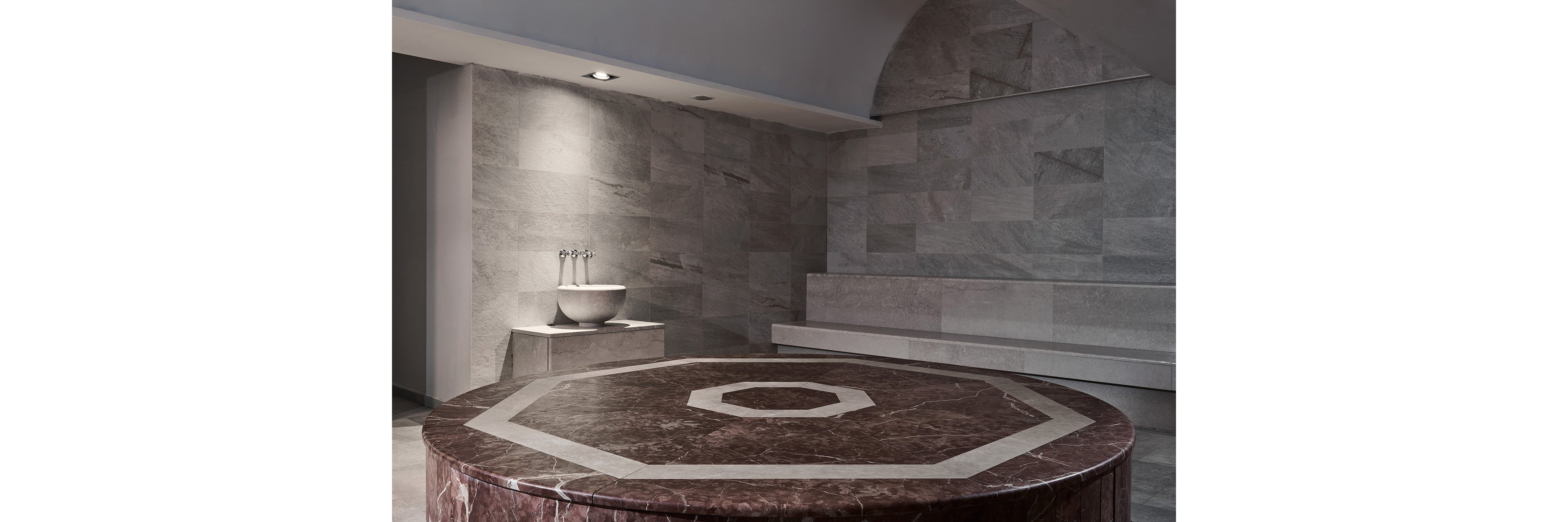 A contemporary Turkish bath interior features a large round marble platform with geometric inlay at the center. The room is clad in smooth gray stone tiles, creating a clean and minimalist spa atmosphere. A stone basin with metal taps stands against the wall, while a long bench provides seating along the side. Recessed ceiling lighting softly illuminates the textured surfaces and architectural details.