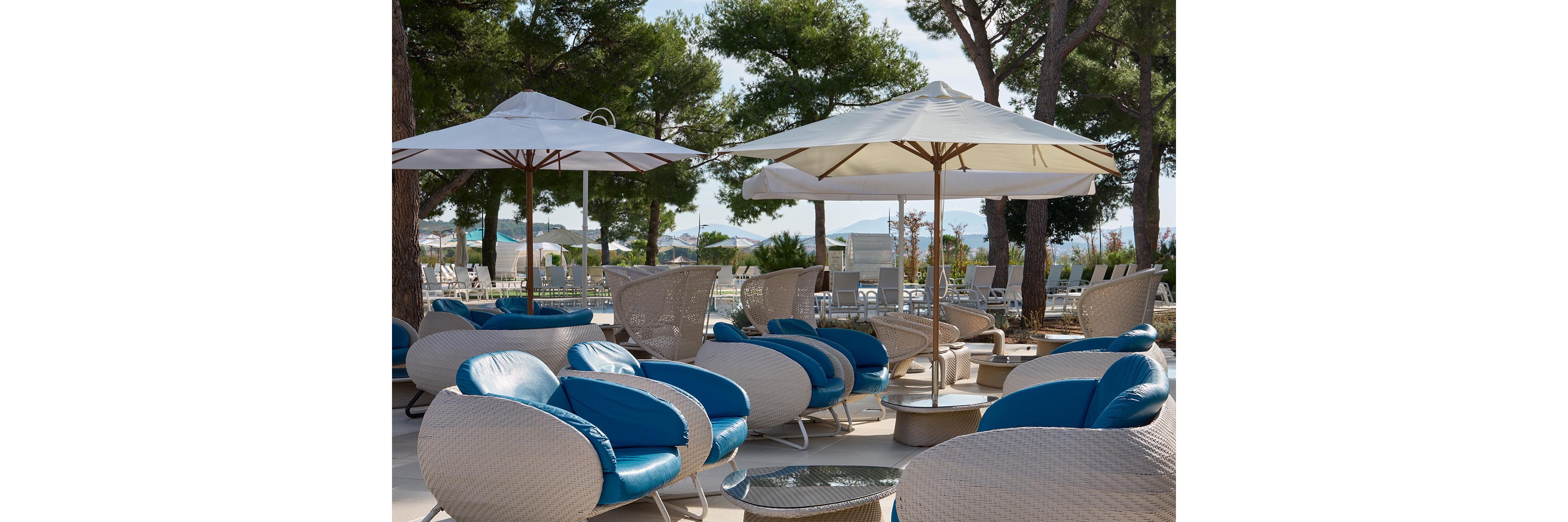 An outdoor resort seating area features modern wicker lounge chairs with blue cushions. Large white patio umbrellas provide shade among tall pine trees. In the background, additional sun loungers and tables are arranged near a pool or terrace. The scene suggests a relaxed vacation atmosphere in a warm, sunny climate.