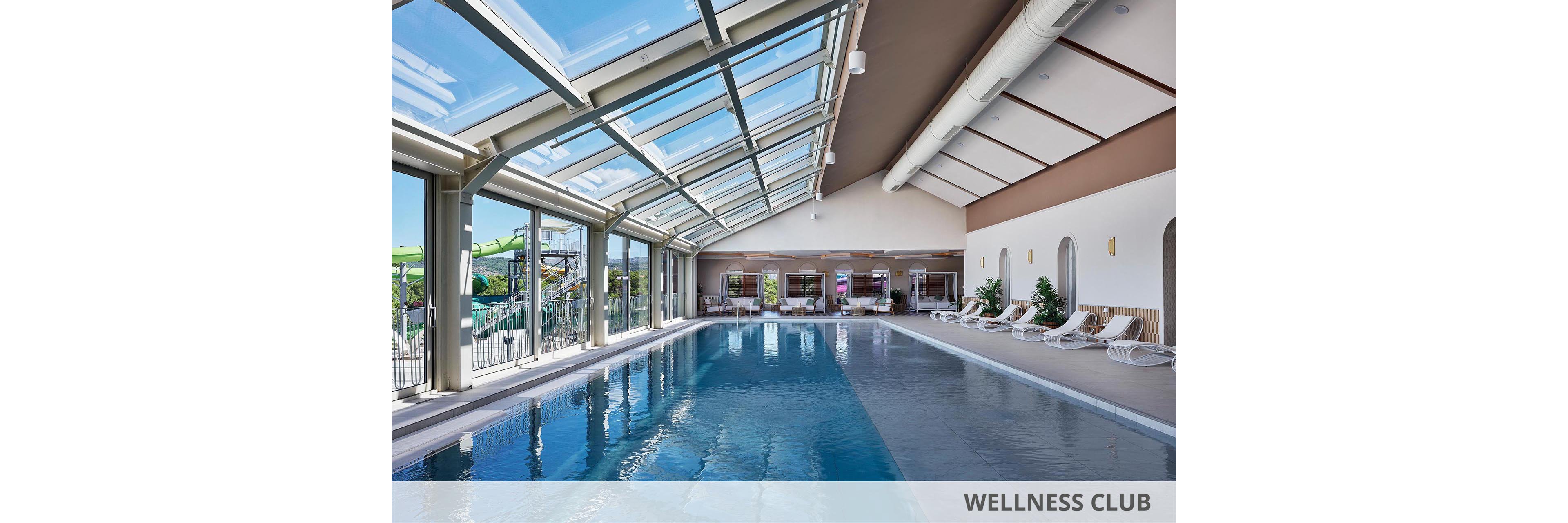 Spacious indoor swimming pool with clear blue water is housed beneath a large glass skylight roof. Sun loungers line the far wall, creating a relaxing resort atmosphere. Floor-to-ceiling windows reveal an adjacent outdoor water park area. The bright, clean interior features neutral tones and modern architectural details, emphasizing comfort and leisure.
