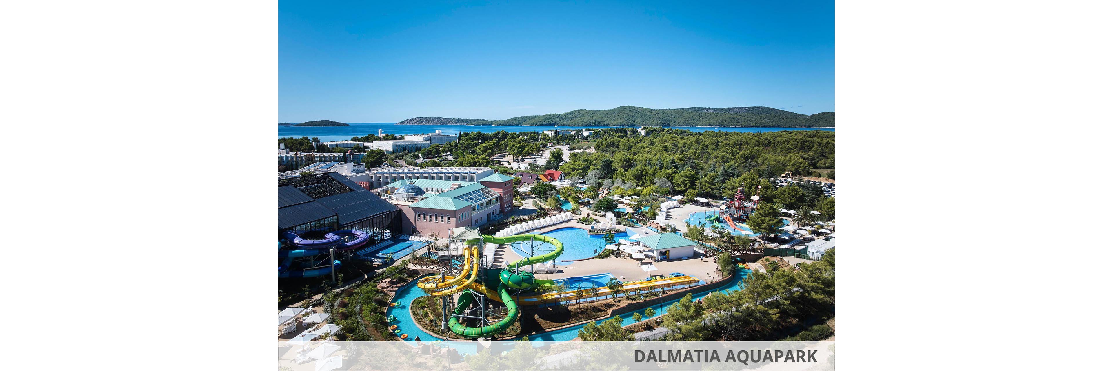 An expansive water park resort is captured from an elevated viewpoint near the coast. Multiple winding water slides, lazy rivers, and pools are surrounded by resort buildings and sun loungers. Dense green trees frame the complex, extending toward the shoreline and distant hills. The calm blue sea and clear sky emphasize a sunny, summer vacation atmosphere.