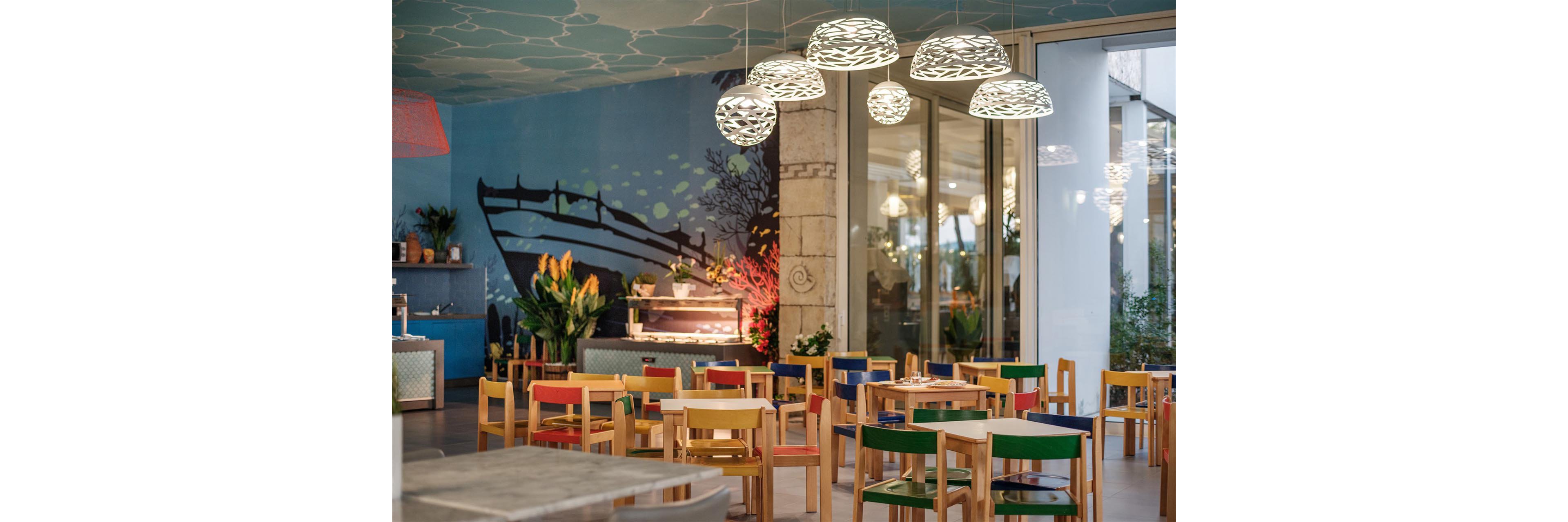 A bright café interior features small wooden tables and chairs with colorful backs and seats. Artistic pendant lights hang from the ceiling, casting a warm glow over the dining area. A large wall mural with an abstract boat and underwater theme decorates the background. Green plants and flowers add natural accents near the counter and along the walls, while large windows open to an outdoor space.