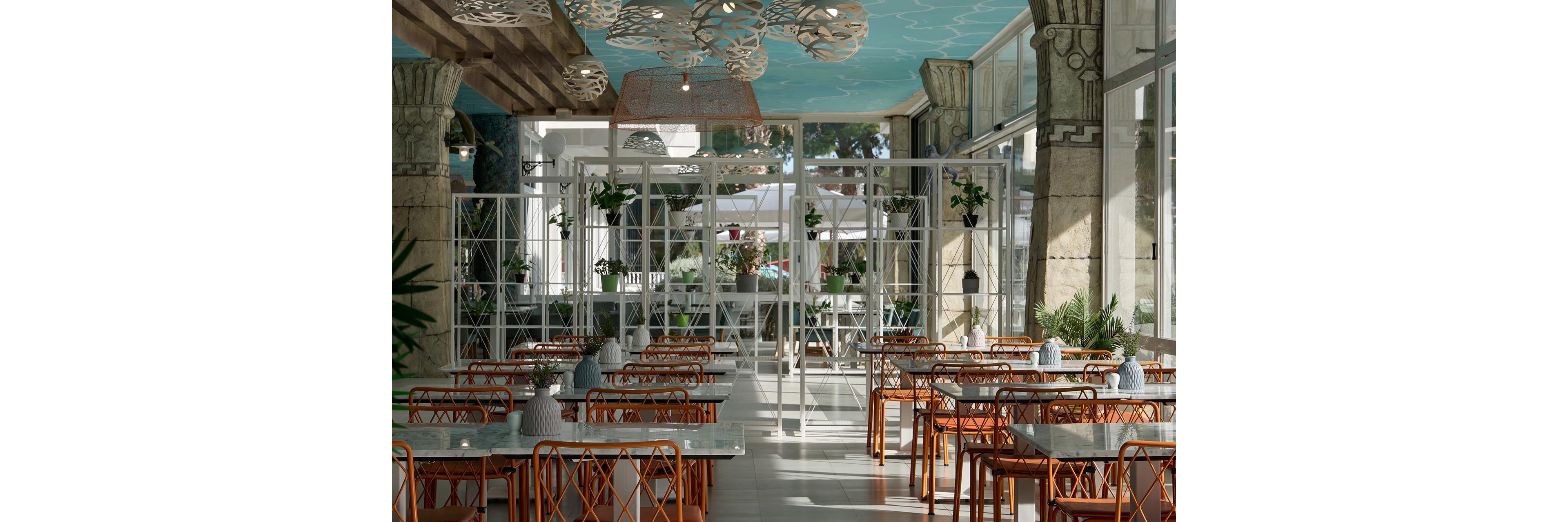 A bright, contemporary restaurant interior features rows of empty tables with orange metal chairs and light-colored tabletops. Large windows line the space, allowing natural light to flood in and offering views of greenery outside. Decorative ceiling fixtures and patterned columns add an artistic touch to the airy setting. Potted plants on shelves and tables contribute to a fresh, inviting atmosphere suitable for dining or casual gatherings.