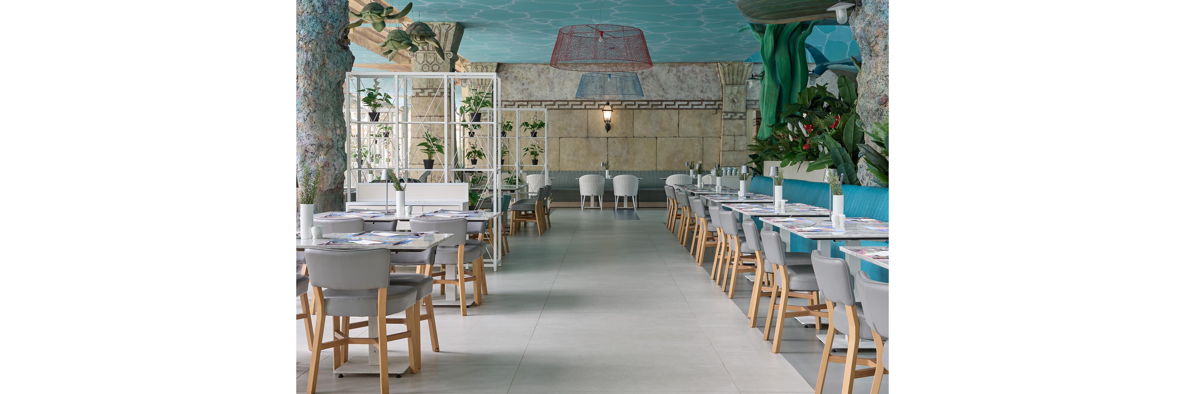 A spacious restaurant interior features rows of neatly arranged tables and gray upholstered chairs. The setting has a coastal and underwater theme, with blue ceiling details, sea-inspired decor, and lush green plants. Soft lighting and neutral floor tiles create a calm, inviting atmosphere. The space appears ready for guests, with place settings laid out along both sides of the room.