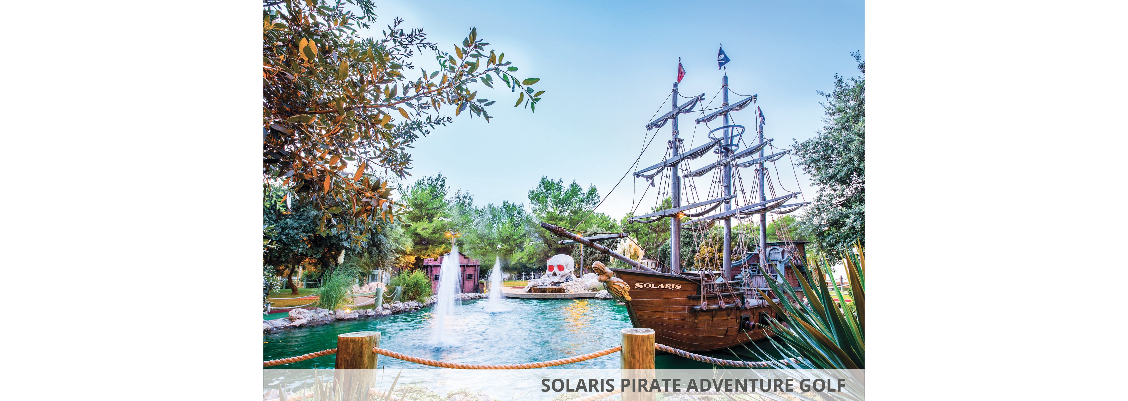 A pirate-themed mini golf course featuring a detailed ship replica with 'Solaris' branding. The setting includes fountains, lush greenery, and a skull-shaped structure, creating a playful and adventurous atmosphere. The scene is bright and colorful, with clear skies enhancing the vibrant visuals.