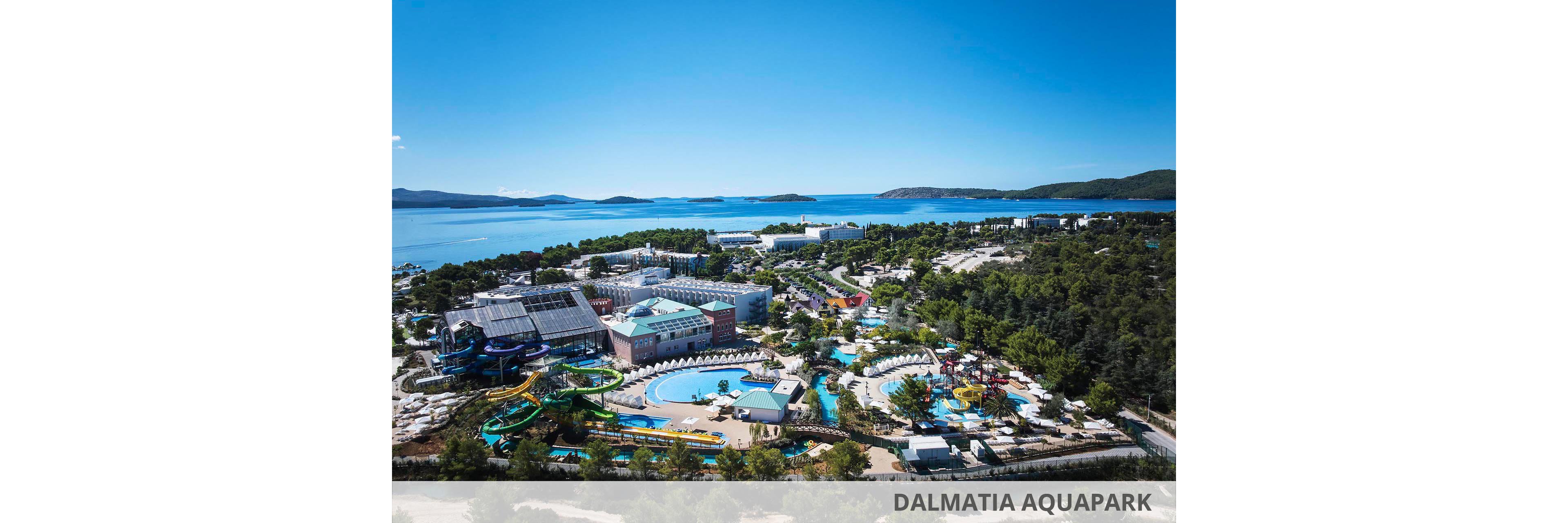A large coastal resort complex with an expansive water park is seen from an elevated viewpoint. Multiple pools, winding water slides, and sun loungers are surrounded by trees and low-rise buildings. Beyond the resort, a calm blue bay stretches out with small islands and distant hills under a clear sky. The scene suggests a popular summer vacation destination with family-friendly attractions and seaside access.