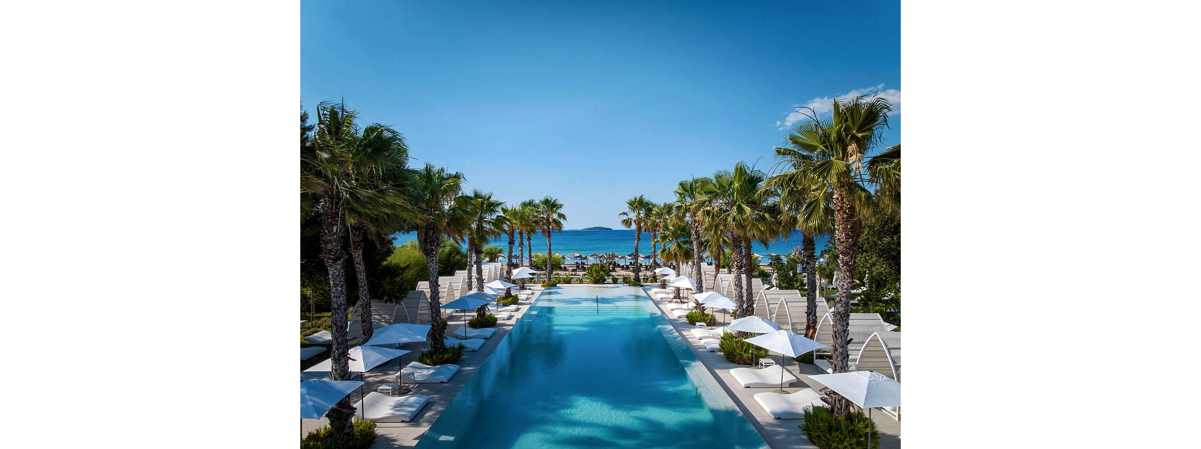 A long, turquoise infinity pool stretches toward the sea, framed symmetrically by tall palm trees. White sun loungers and umbrellas line both sides of the pool, creating a high-end resort atmosphere. In the distance, the blue ocean and a small island are visible under a clear sky. The overall scene feels calm, sunny, and luxurious, ideal for a tropical vacation setting.