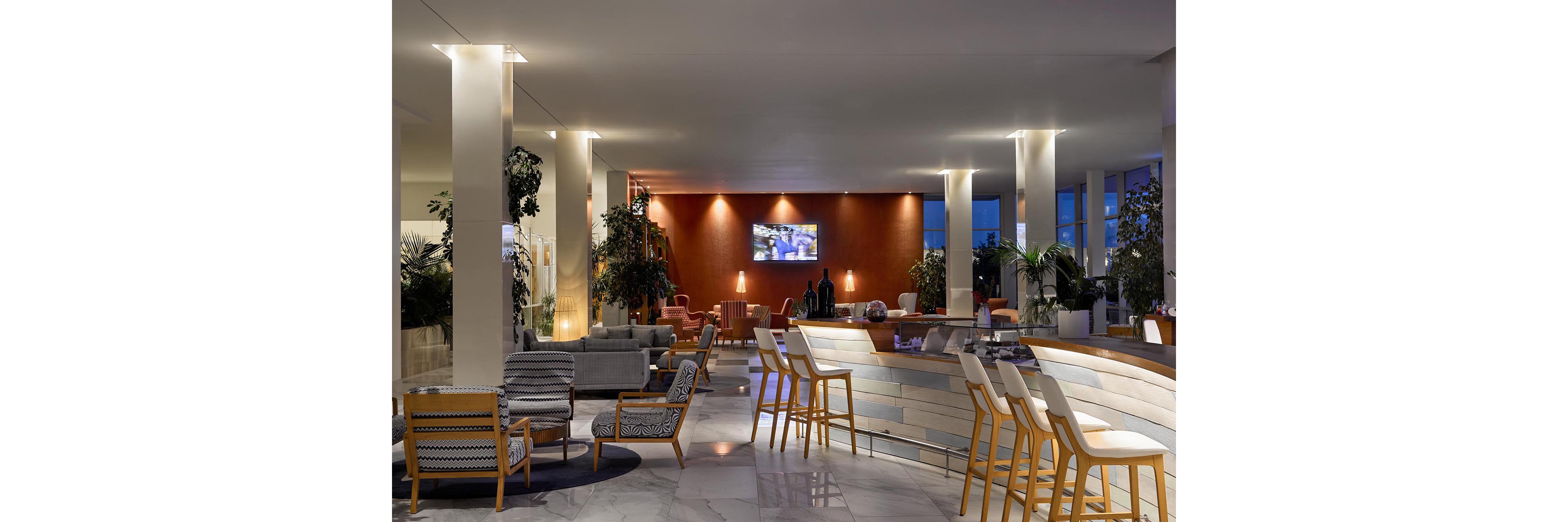 A contemporary hotel lobby features a curved bar with high wooden stools and cushioned seating. Lounge chairs and sofas are arranged in small conversation areas across a polished marble floor. Warm wall lighting and a large television screen create a relaxed, inviting atmosphere. Tall columns and indoor plants add structure and greenery to the spacious interior.