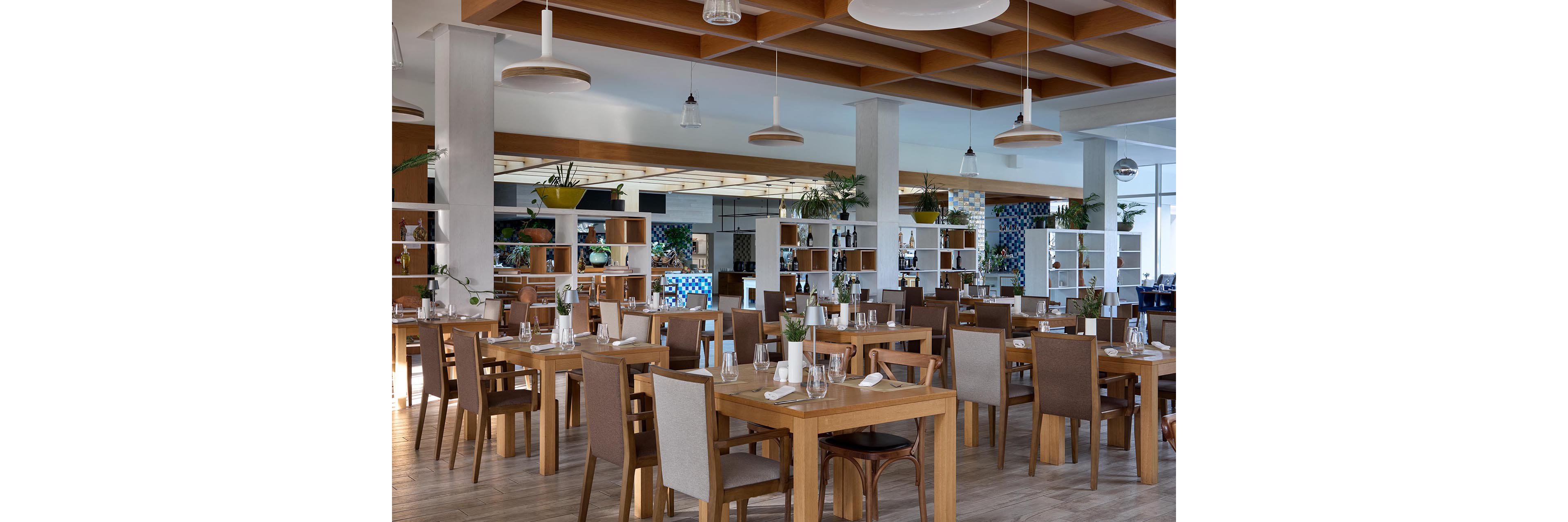 Contemporary restaurant interior featuring neatly arranged wooden tables and upholstered chairs. The dining room is empty, with place settings of glasses, plates, and folded napkins prepared for guests. Large windows and pendant lights create a bright, airy atmosphere, while open shelving with plants and bottles adds decorative interest. The overall design feels clean, modern, and welcoming.