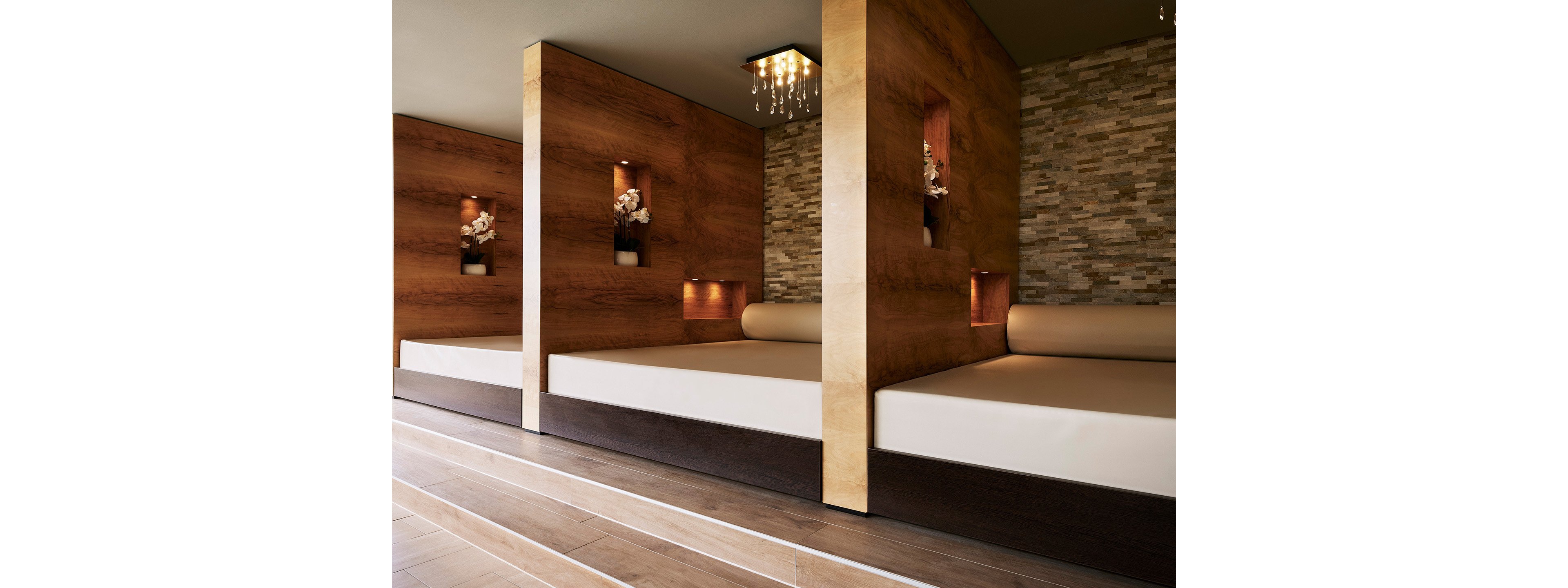 Image of a modern spa interior featuring several large cushioned relaxation beds separated by wooden partitions. Each partition includes recessed niches with decorative orchids and soft accent lighting. The background wall is covered with stacked stone tiles, adding texture and a natural feel. The overall design is clean and contemporary, with warm tones and polished wooden flooring creating a tranquil atmosphere.