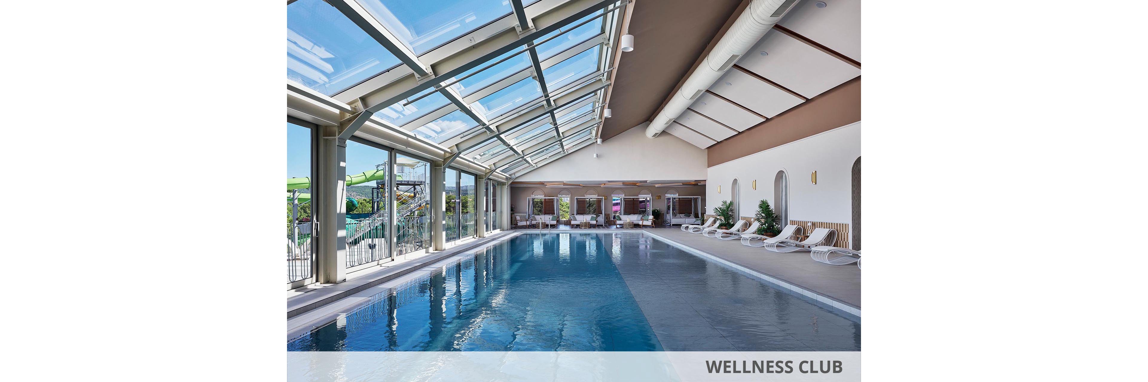 Spacious indoor swimming pool with clear blue water stretches along a sunlit room. Large floor-to-ceiling windows and a glass roof let in abundant natural light and views of an outdoor water park. White lounge chairs line the poolside near potted plants and a seating area in the background. The clean, modern design and calm atmosphere suggest a relaxing hotel or resort wellness space.