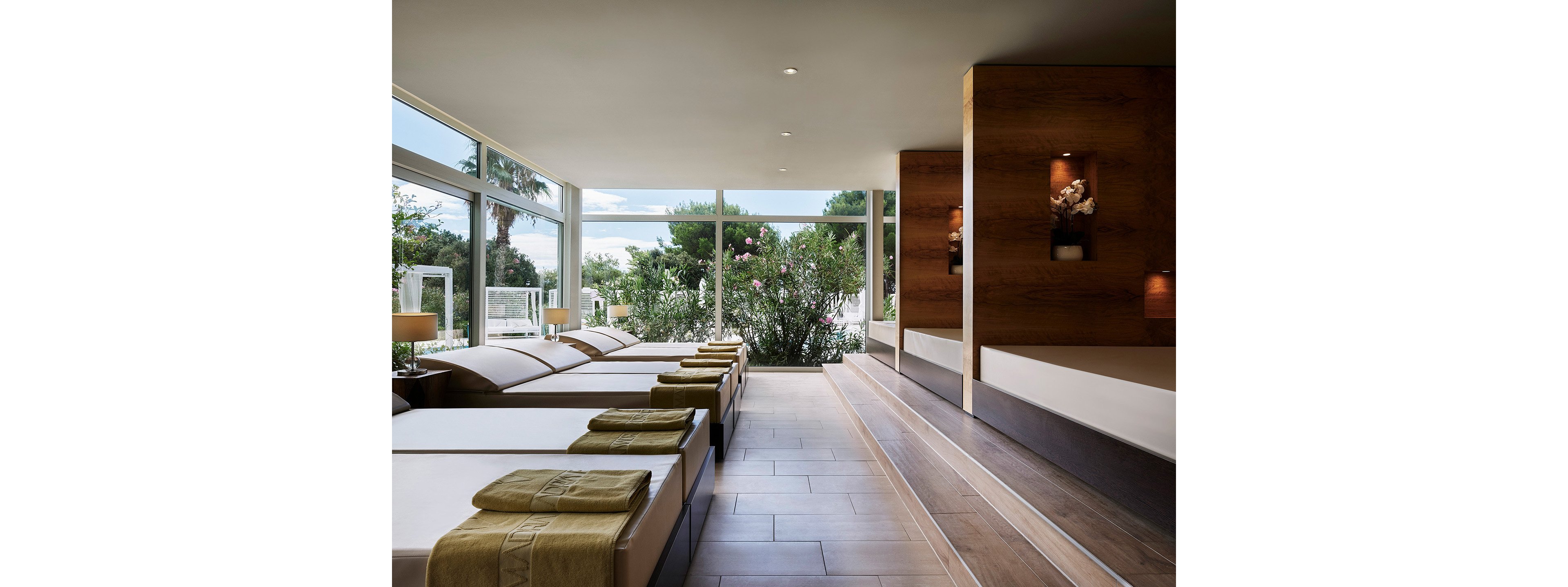 A contemporary spa relaxation room features several cushioned loungers neatly arranged in a row. Large floor-to-ceiling windows reveal lush greenery and flowering bushes outside, filling the space with natural light. Soft neutral tones, wooden paneling, and subtle decorative lighting create a calm and luxurious atmosphere. Folded towels on each lounger suggest a prepared, welcoming environment for guests.
